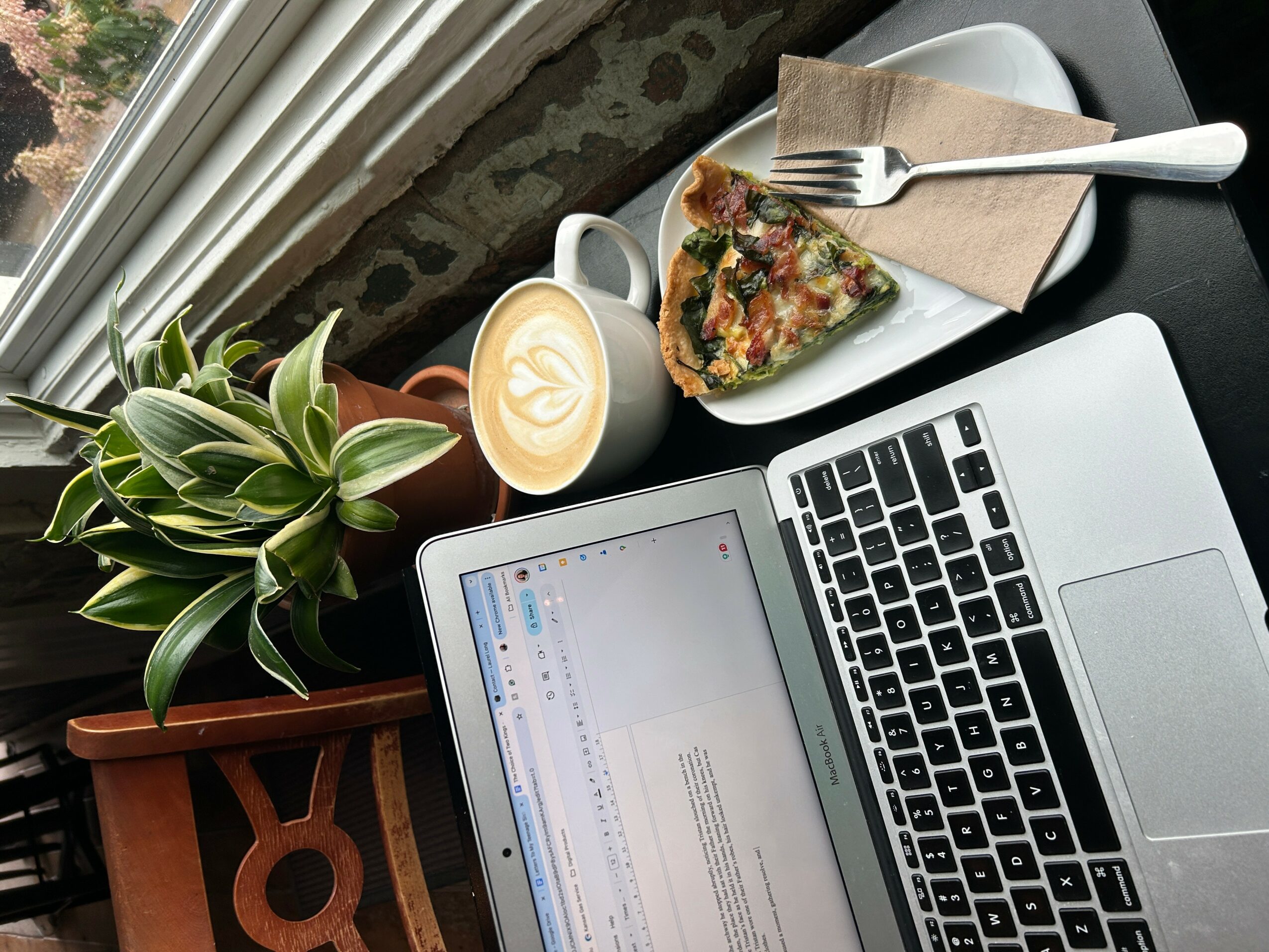 Laptop, plant, coffee, and pizza on a table near a window.