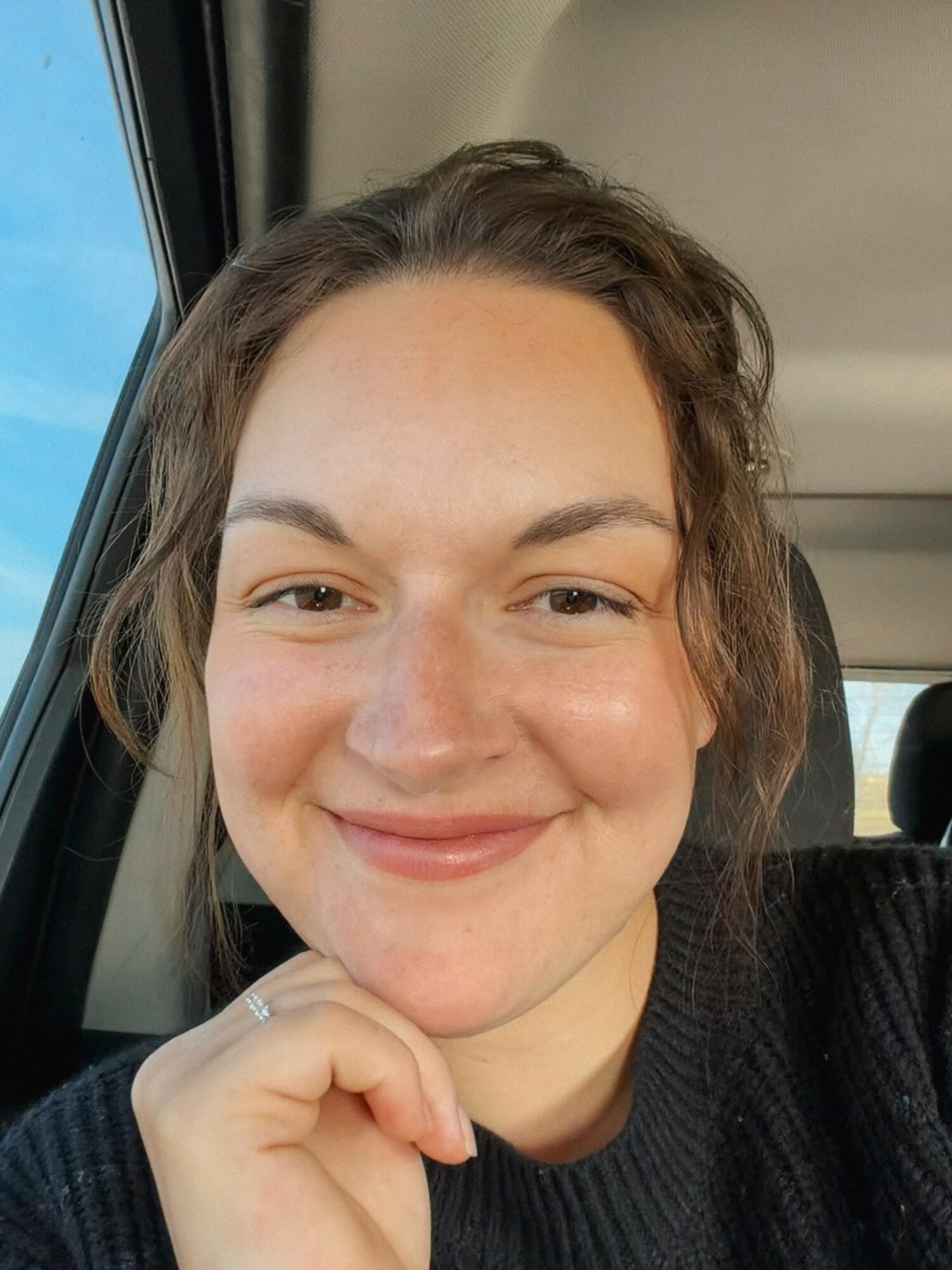 Smiling woman with brown hair in a vehicle, resting her chin on her hand, wearing a black top.