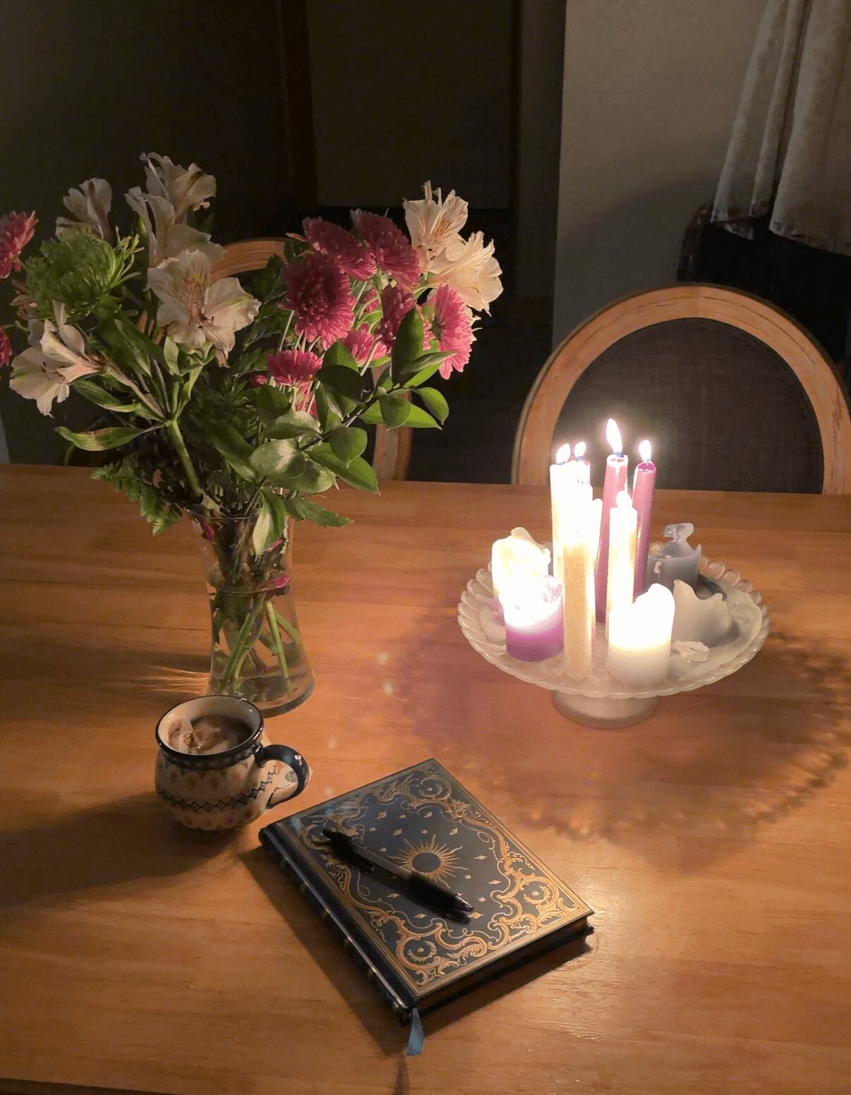Vase with pink and white flowers, lit candles on a plate, closed notebook with pen, on wooden table, dim lighting.