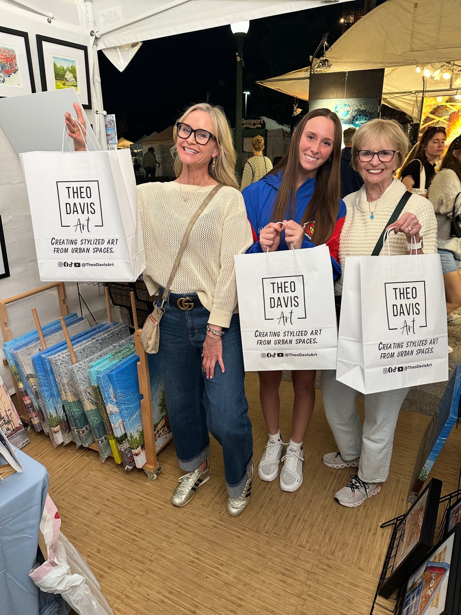 Three women stand together at a book signing event, holding shopping bags, smiling, with books and displays in the background.