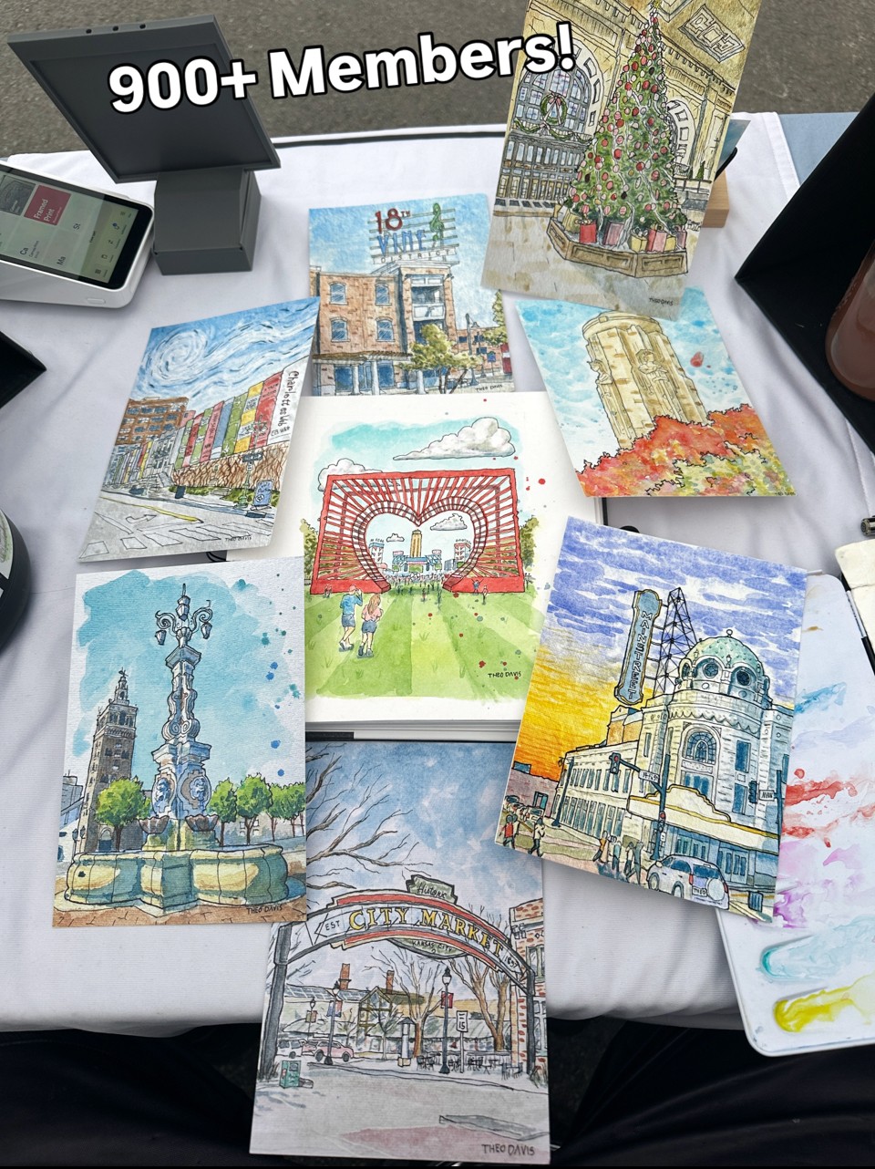 Collection of colorful cityscape paintings on a table with a sign reading '900+ Members!', various objects around.