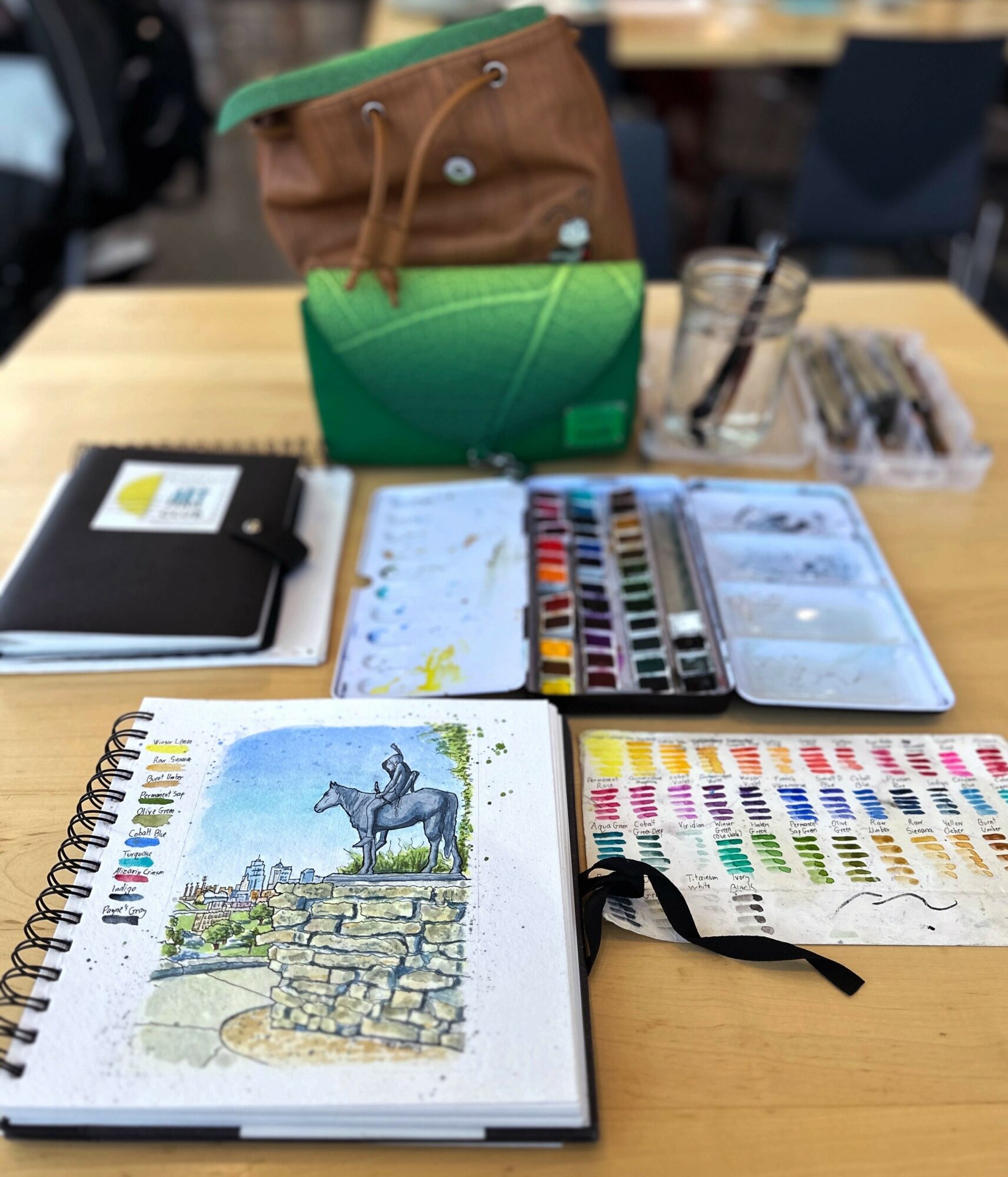 Art supplies on a wooden table, including watercolor paints, a sketchbook with a horse drawing, and a green and brown bag.