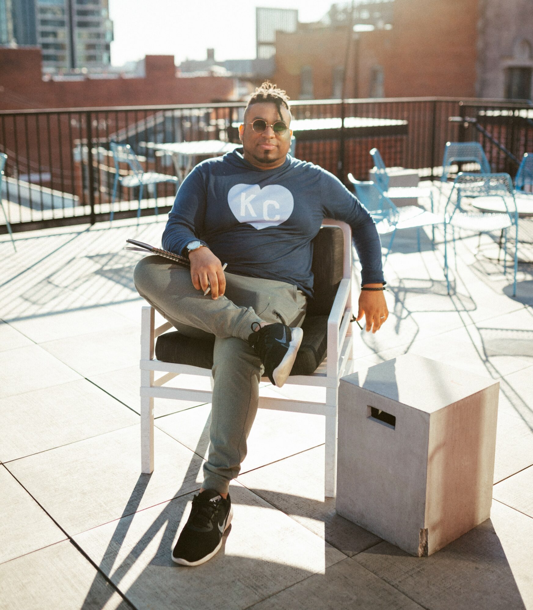 Man sitting on a chair on a rooftop terrace with outdoor furniture, sunlight, and city buildings in background.