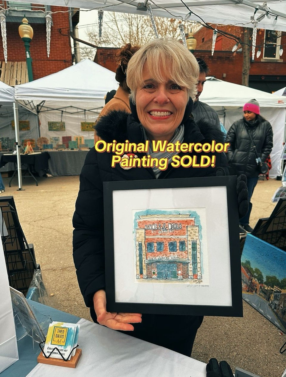 Woman holding framed watercolor painting outdoors at market, smiling, with tents and people in background.
