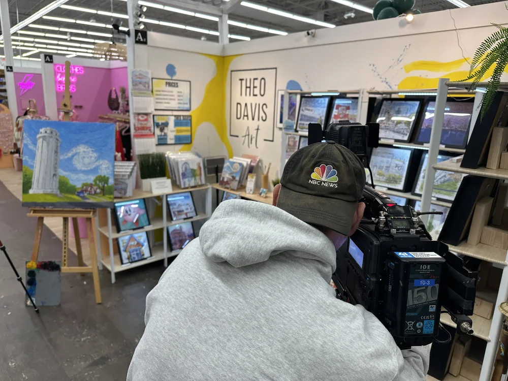 Person wearing a black cap and gray hoodie working with electronic equipment at an art booth, with books and artwork displayed in the background.