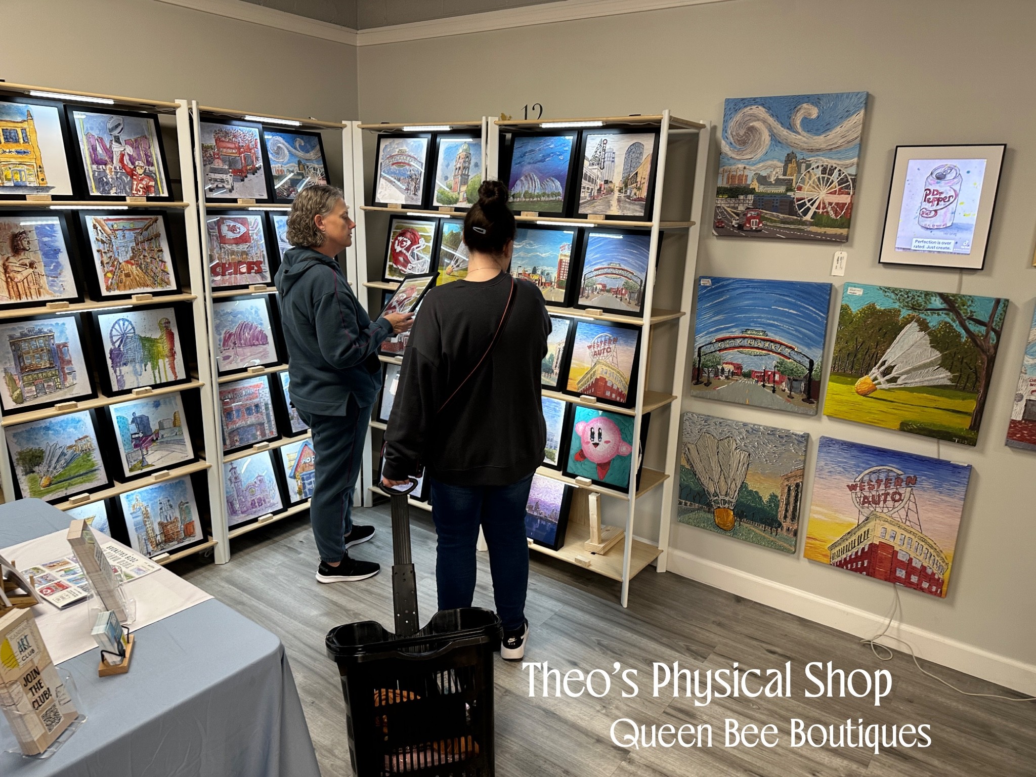 Three people browse bookshelves in a bookstore with colorful artwork on the wall, and a table with items on the left.