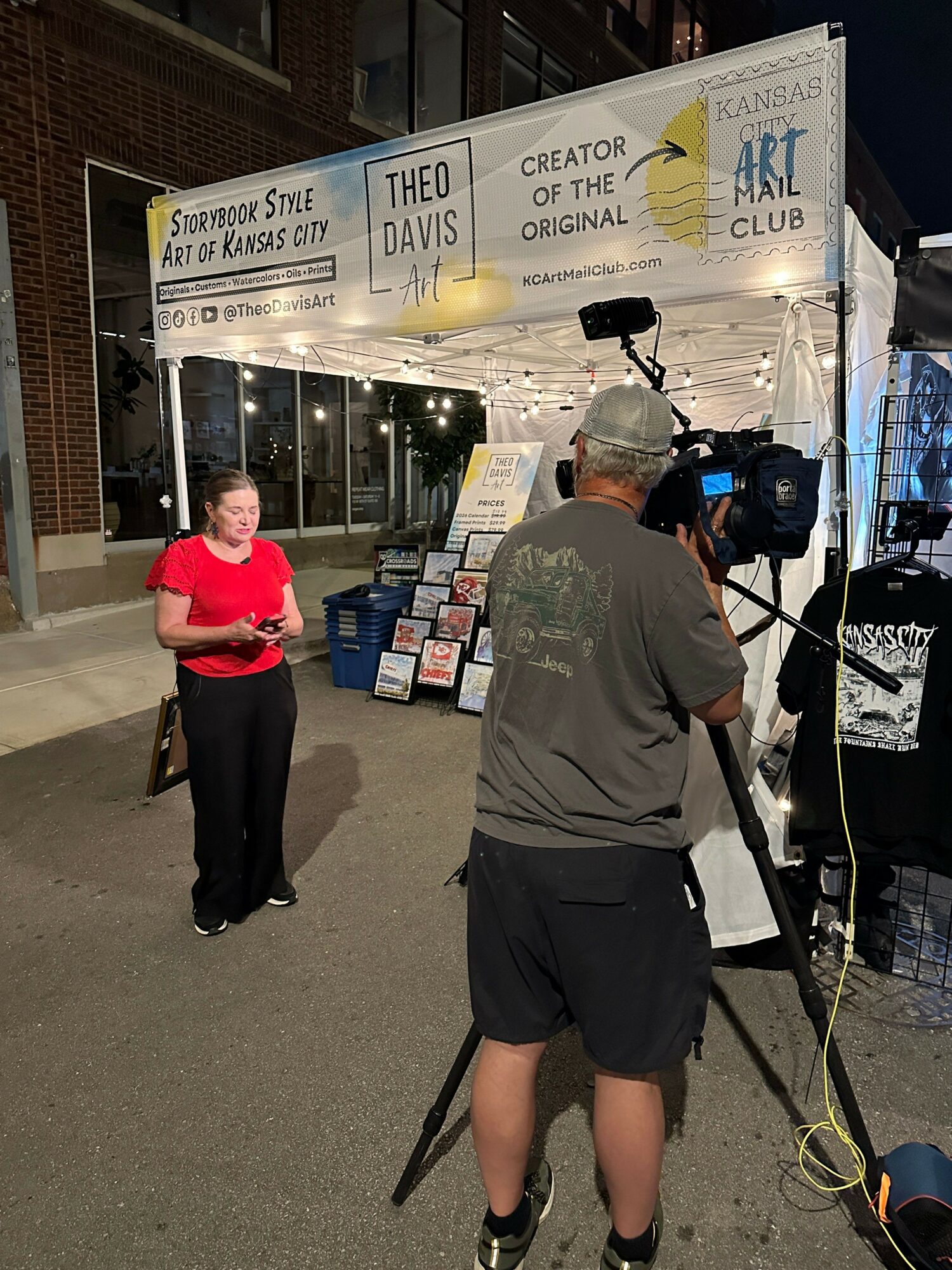 Person in gray shirt and shorts filming at an event booth with a camera, another person in red shirt nearby, booth signage visible.