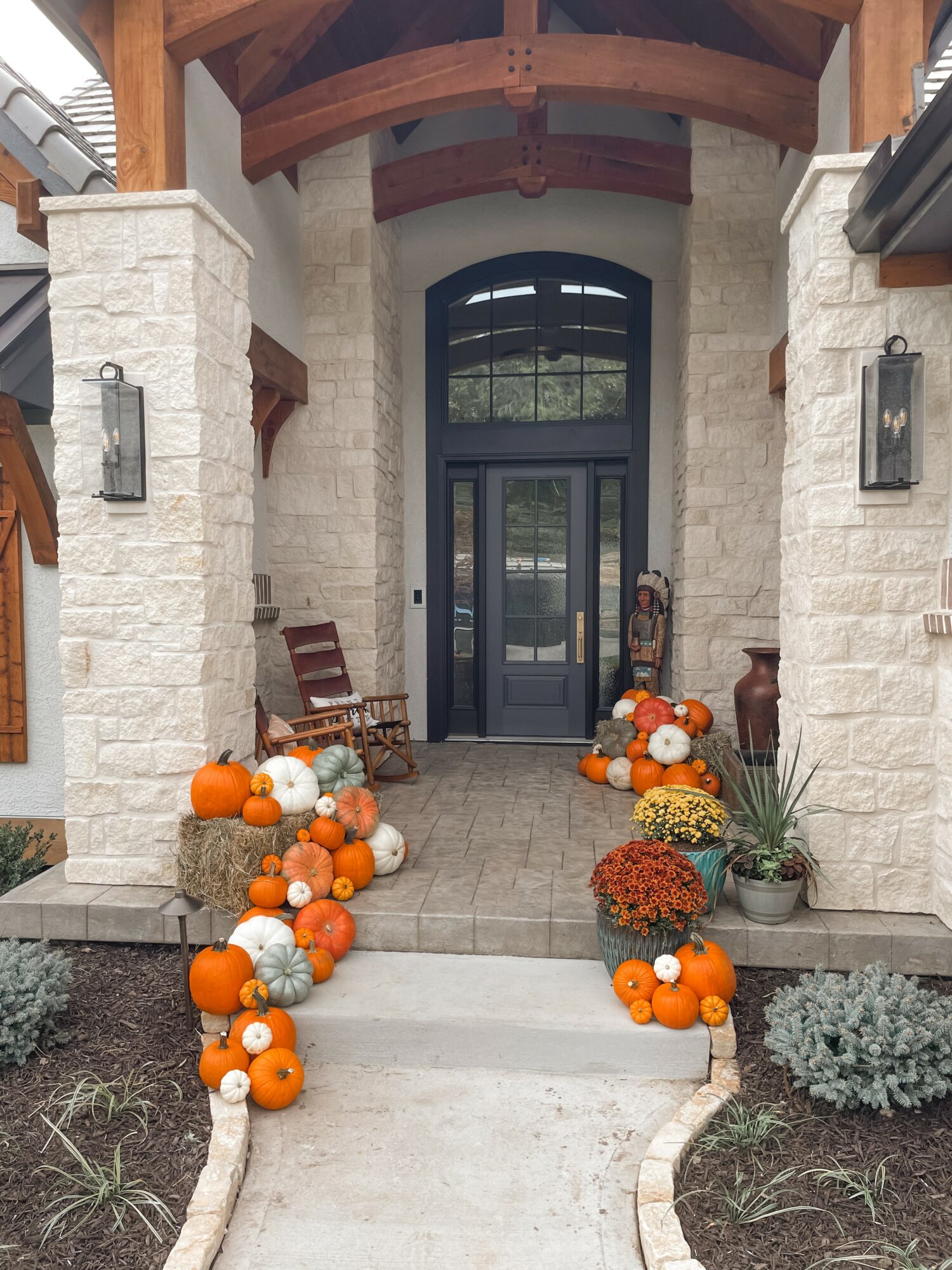 Front entrance with pumpkins and flowers on steps, stone walls, wooden accents, and glass door, autumn decoration.