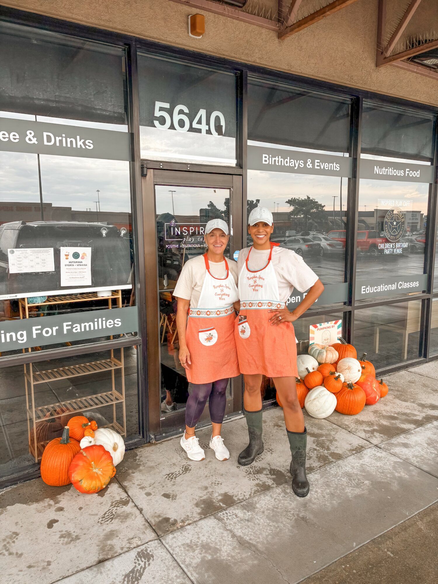 Two people stand outside a store with pumpkins on the ground, wearing aprons and hats, smiling at the camera.