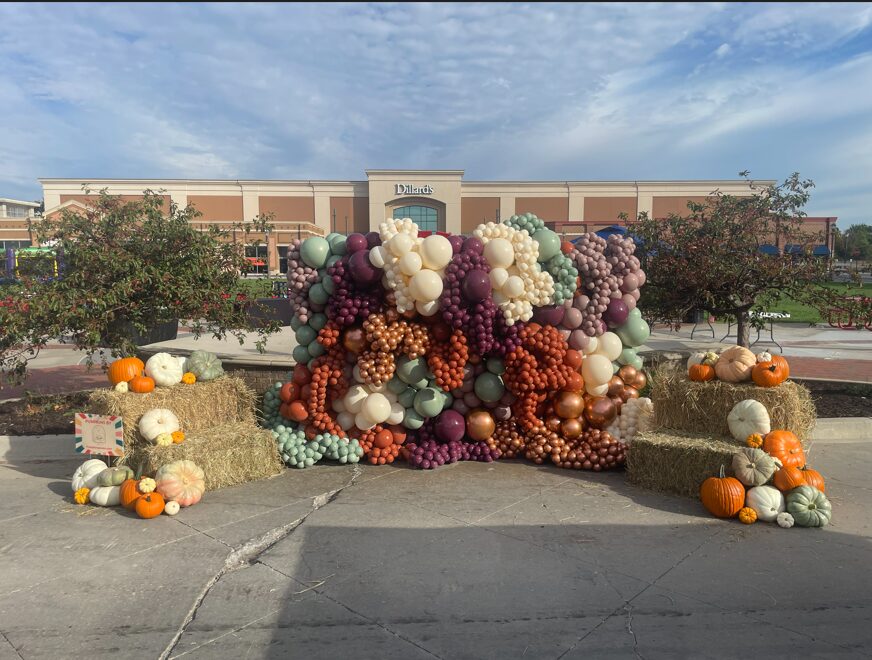 Fall display with pumpkins, hay bales, and balloon arrangement in front of a shopping center, under a partly cloudy sky.