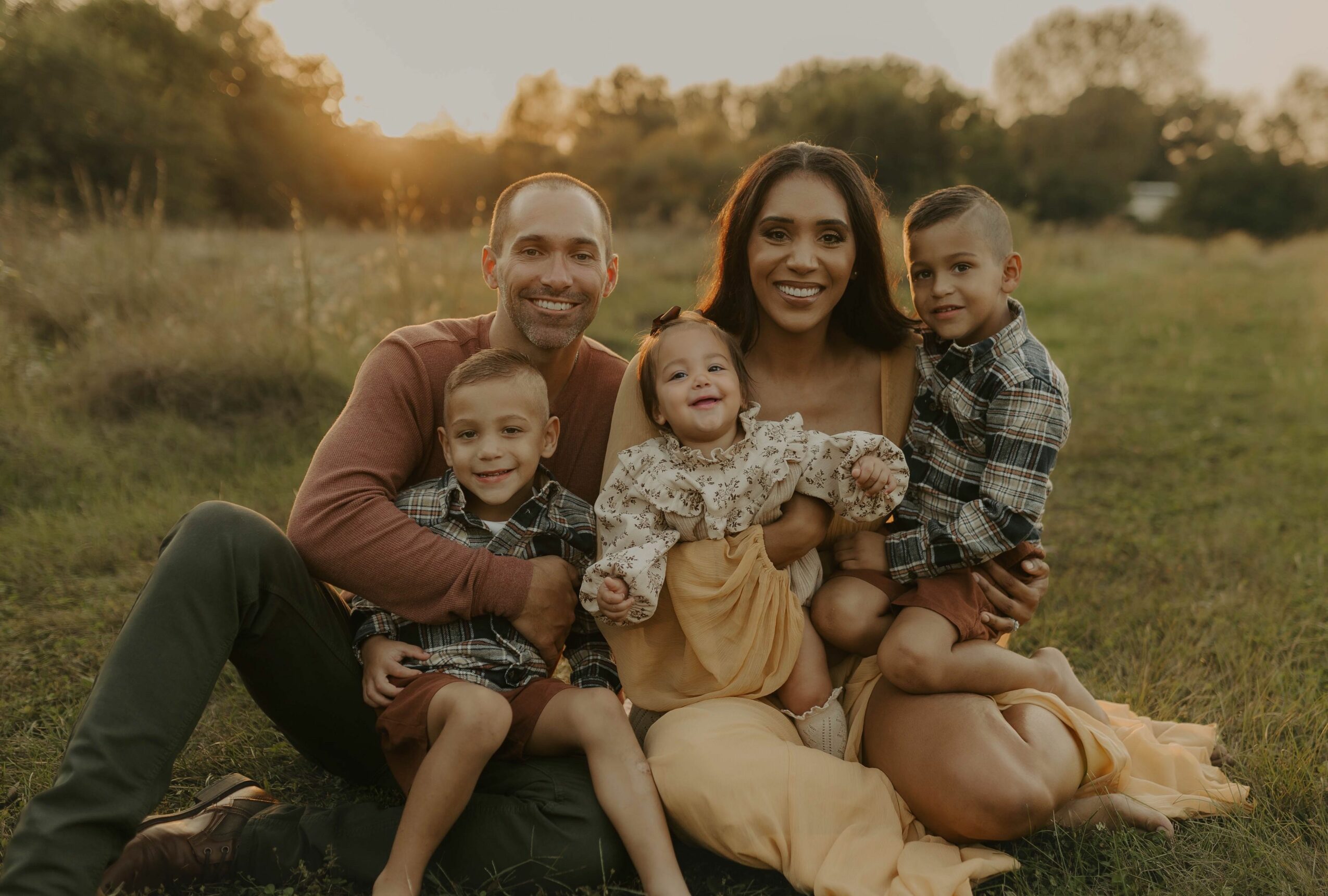 Family of five sitting outdoors on grass during sunset, smiling and enjoying each other's company.