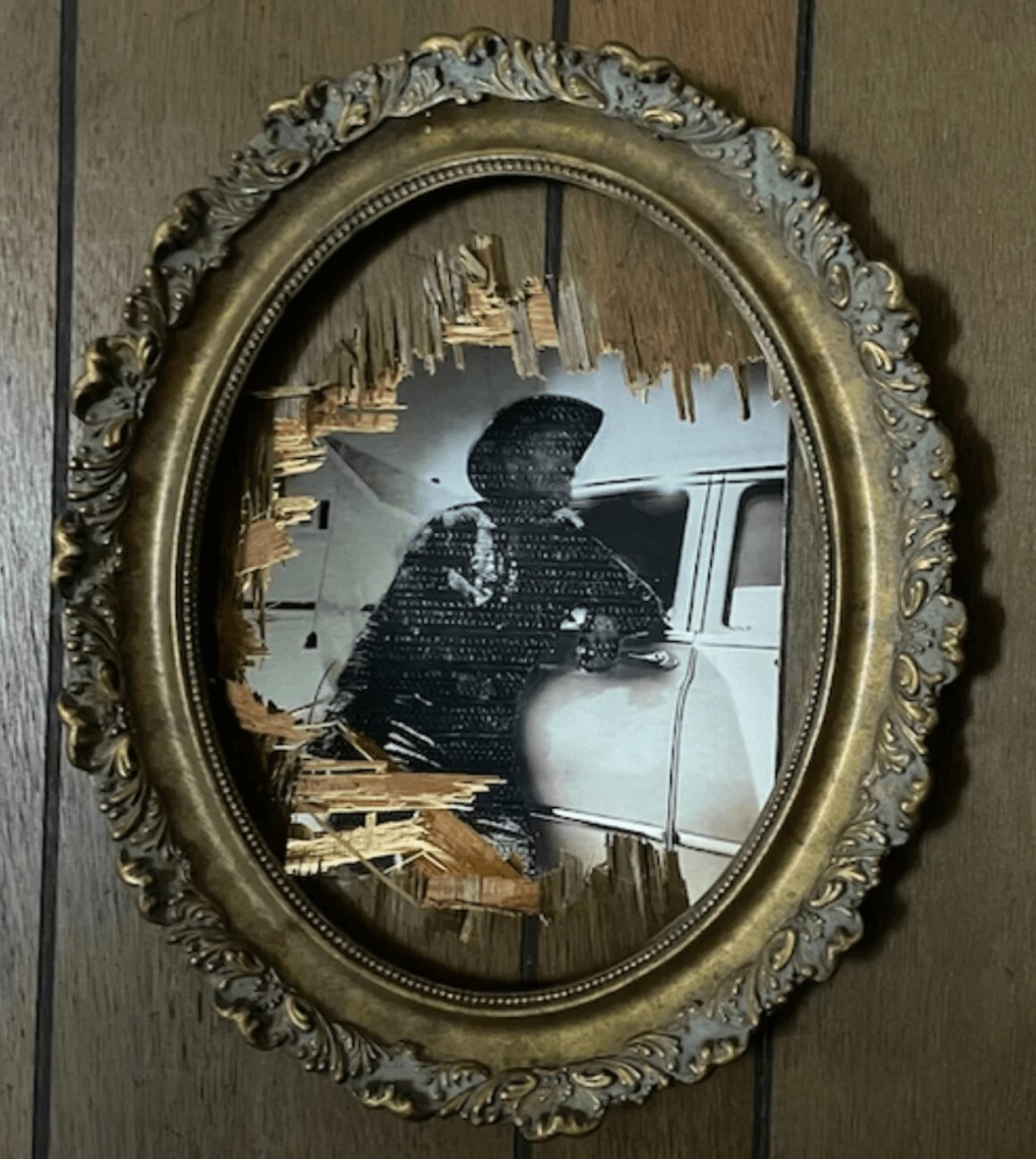 Framed mirror with a damaged reflective surface showing a silhouette of a person and a vehicle, ornate gold frame, wooden background.