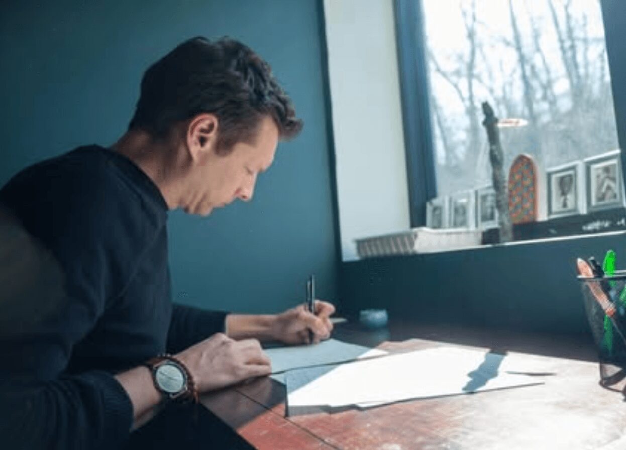 Man sitting at a desk writing on paper near a window with natural light.