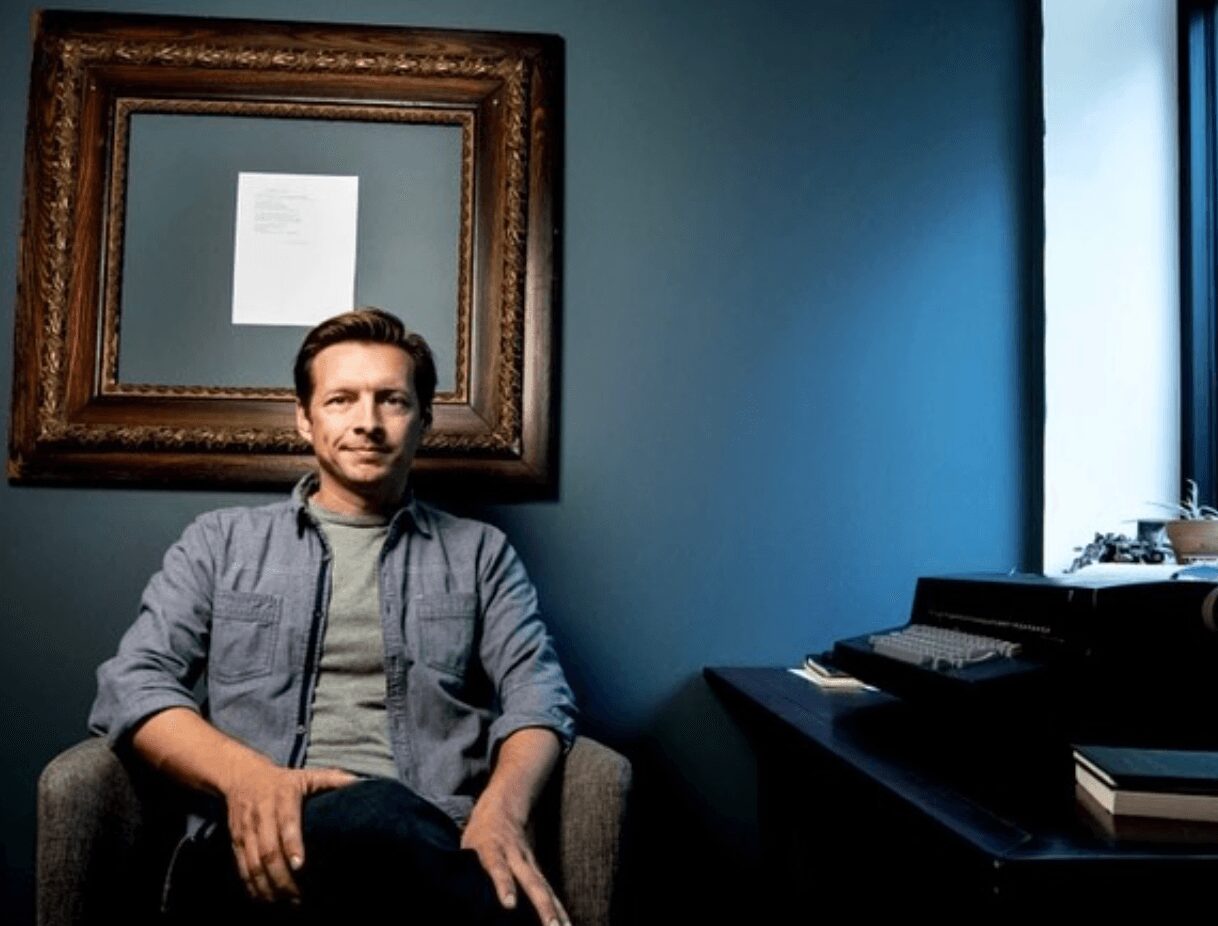 Man sitting in a room with a large framed picture on the wall, near a window with sunlight, and a desk with books.