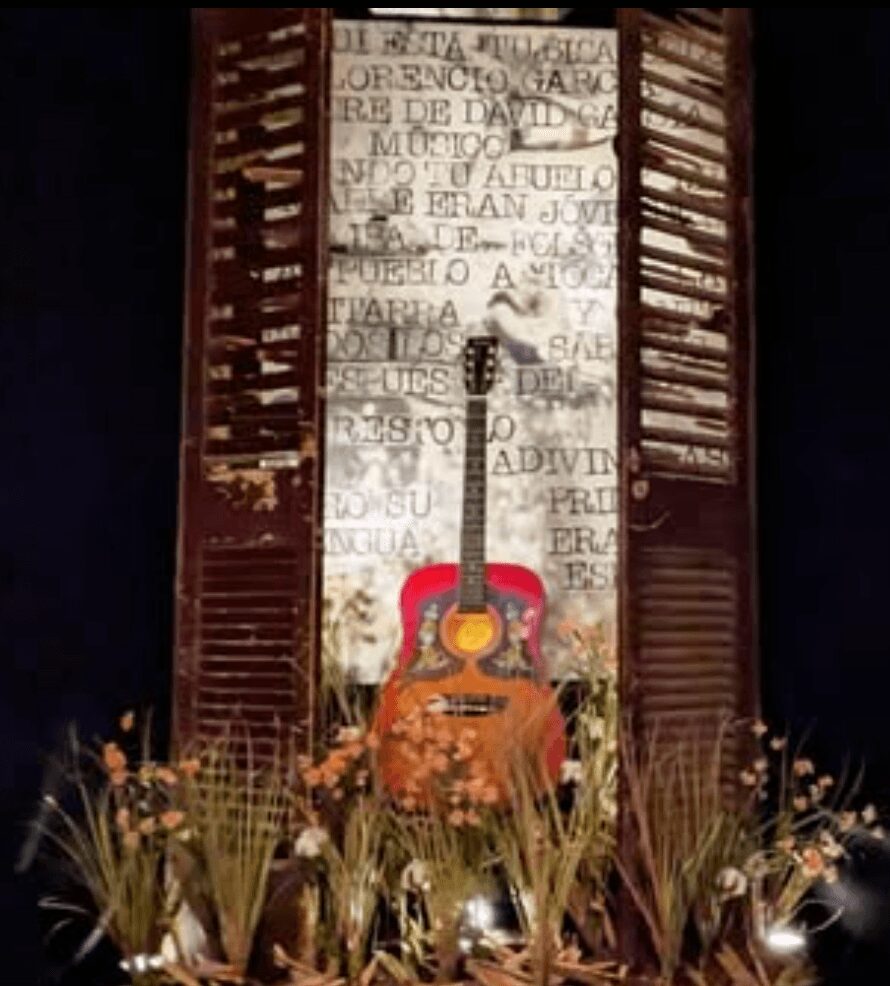 Guitar leaning against a wall with text, flanked by wooden shutters, surrounded by flowers and plants.