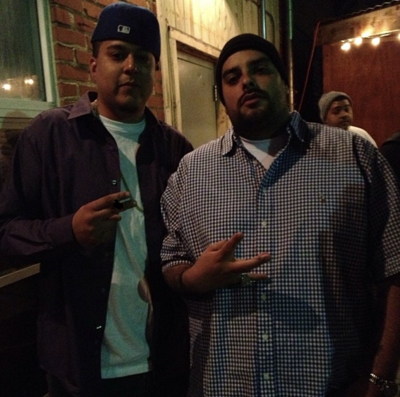 Two men standing outdoors at night, one making a peace sign, with a brick wall and string lights in the background.