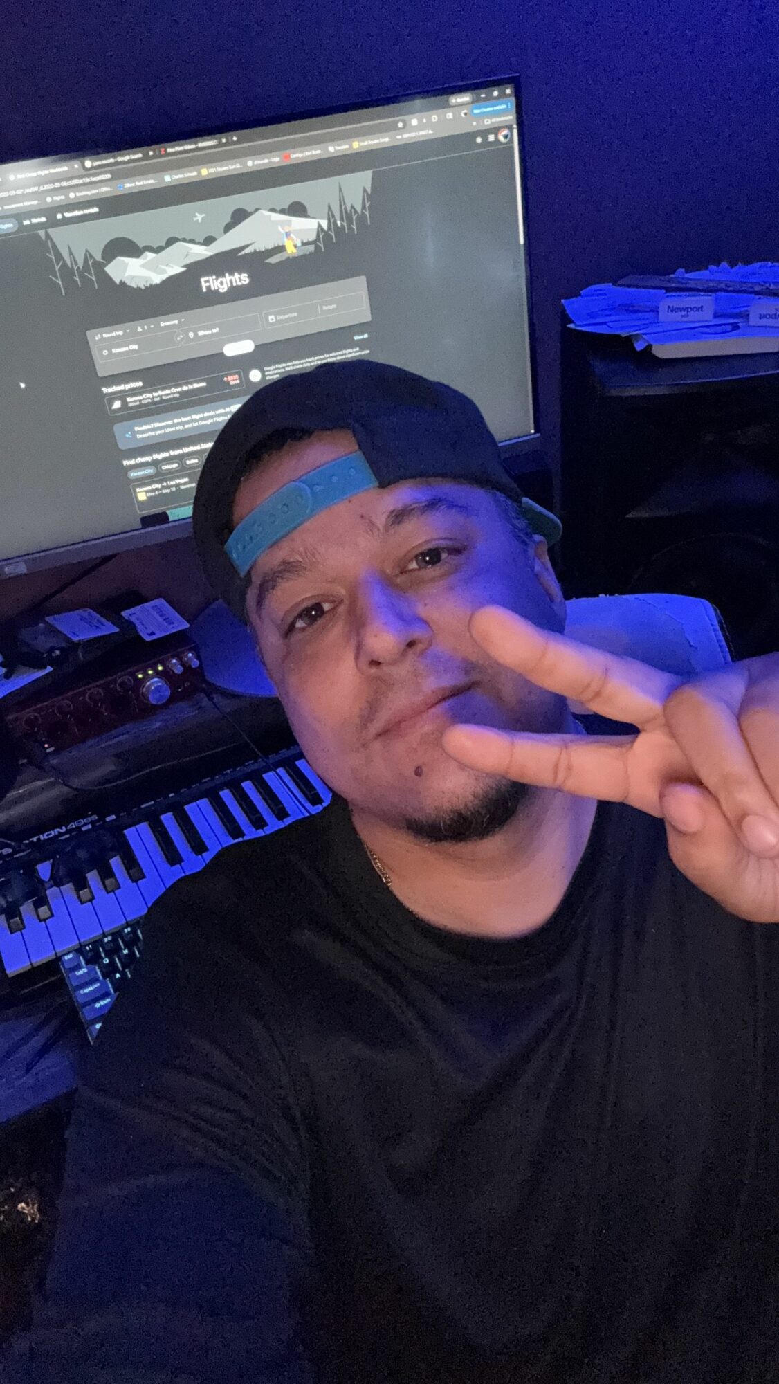Person with a cap making a peace sign near a computer and keyboard, in a dark room with blue lighting.