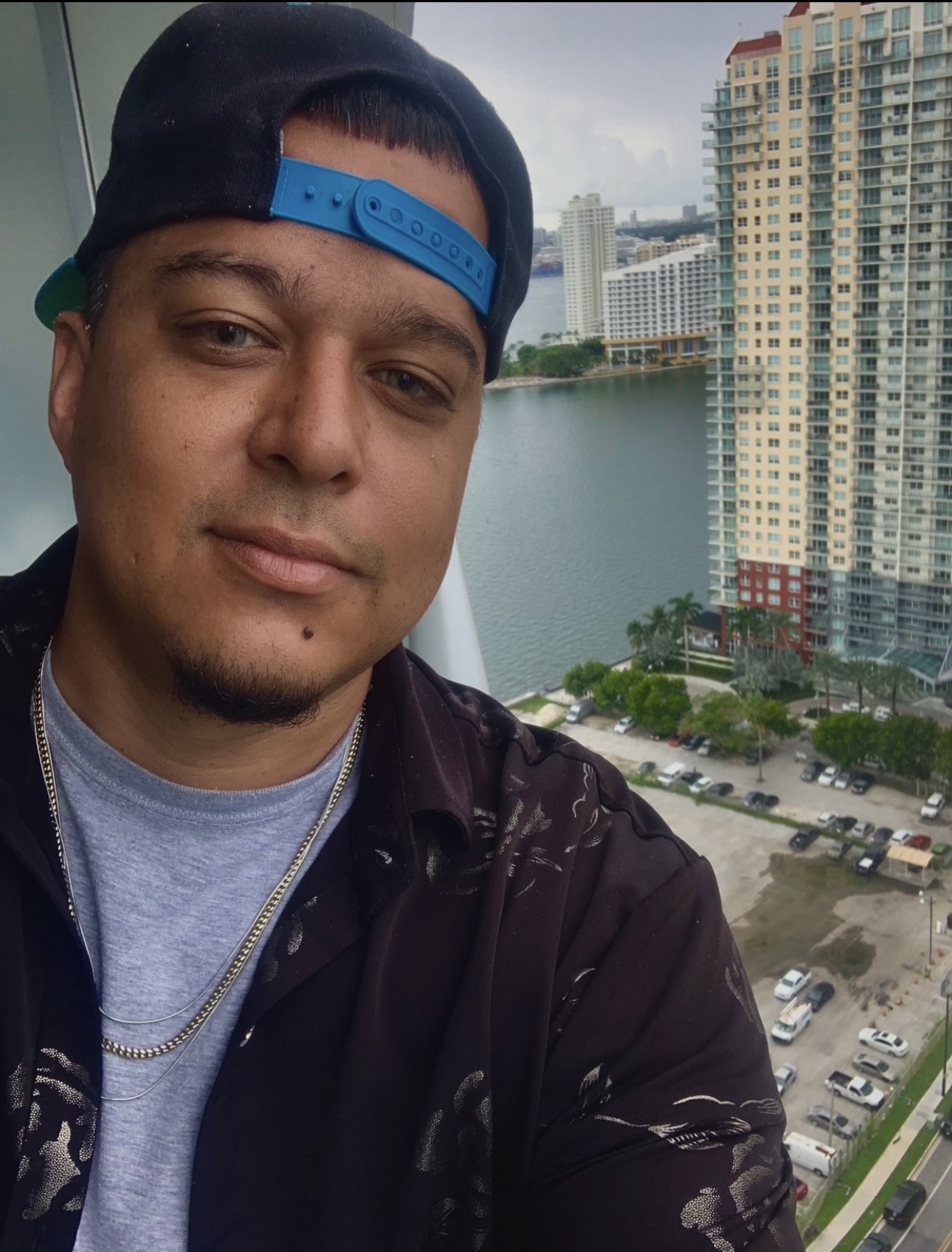 Young man wearing a cap backwards, taking a selfie with a cityscape and river in the background.