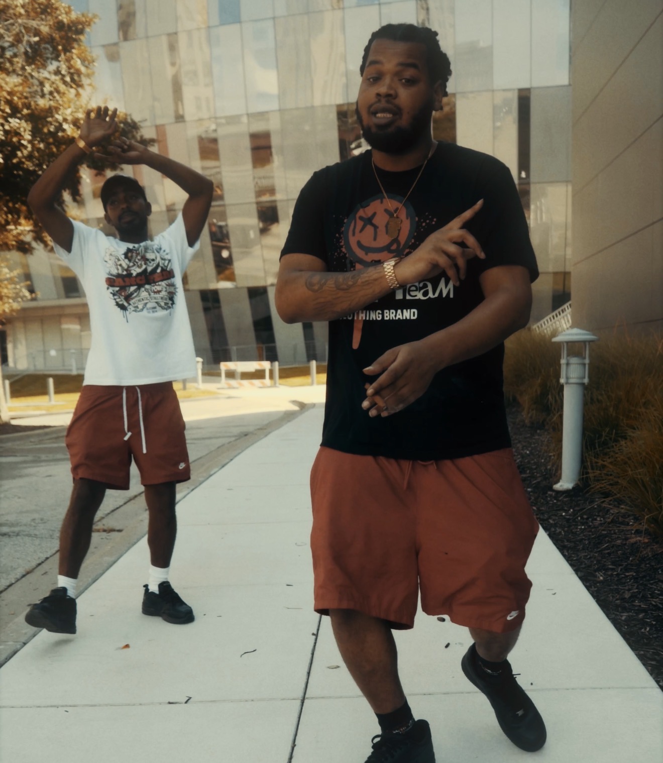 Two men walking outdoors on a sidewalk, one pointing, the other holding a ball, with modern buildings in the background.