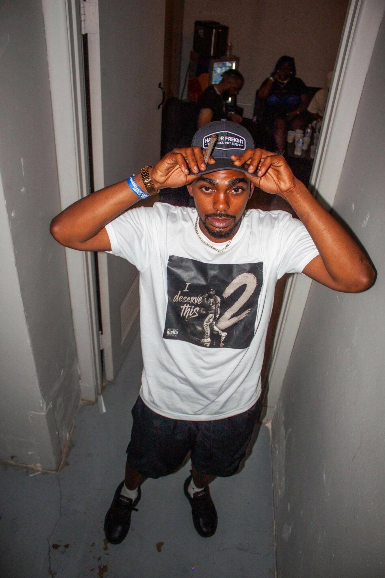 Young man wearing a cap, white t-shirt, and shorts, standing in a narrow hallway, adjusting his cap, with a mirror reflecting others in the background.