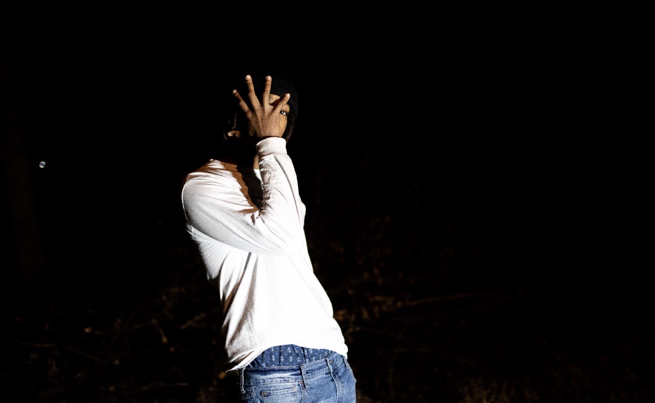 Person standing outdoors at night, covering face with hand, wearing a white long-sleeve shirt and jeans.