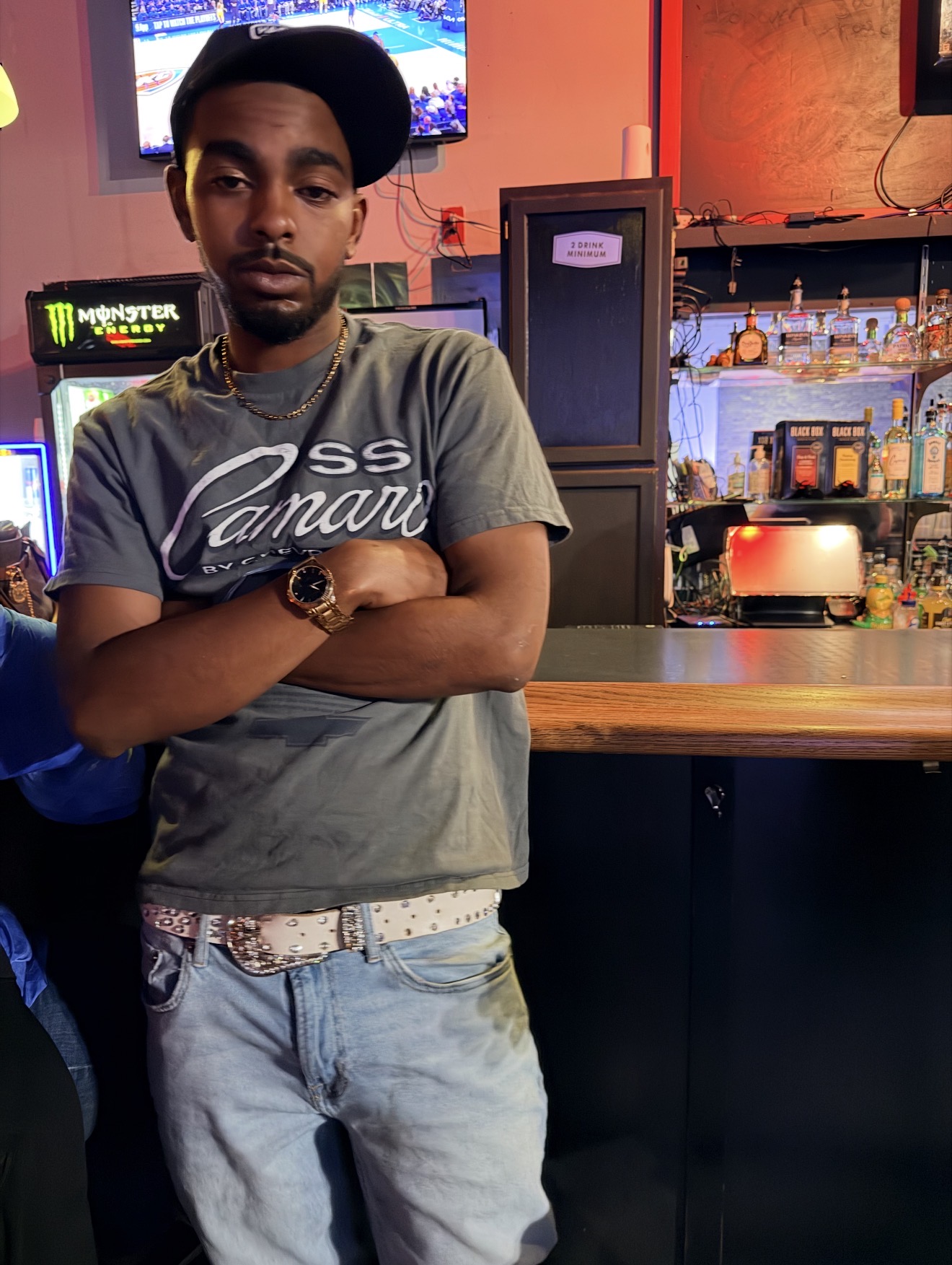Young man with crossed arms wearing a cap and casual clothes in a bar or restaurant setting.