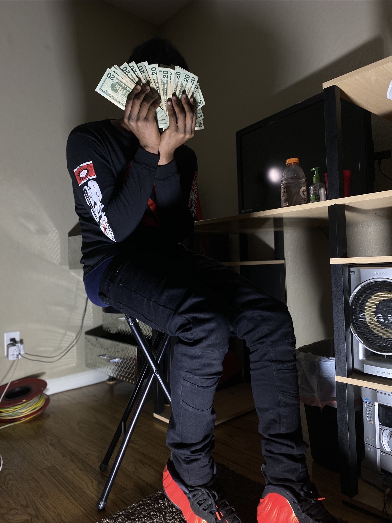 Person sitting on a chair holding cash in front of their face, in a room with a desk and electronic equipment.