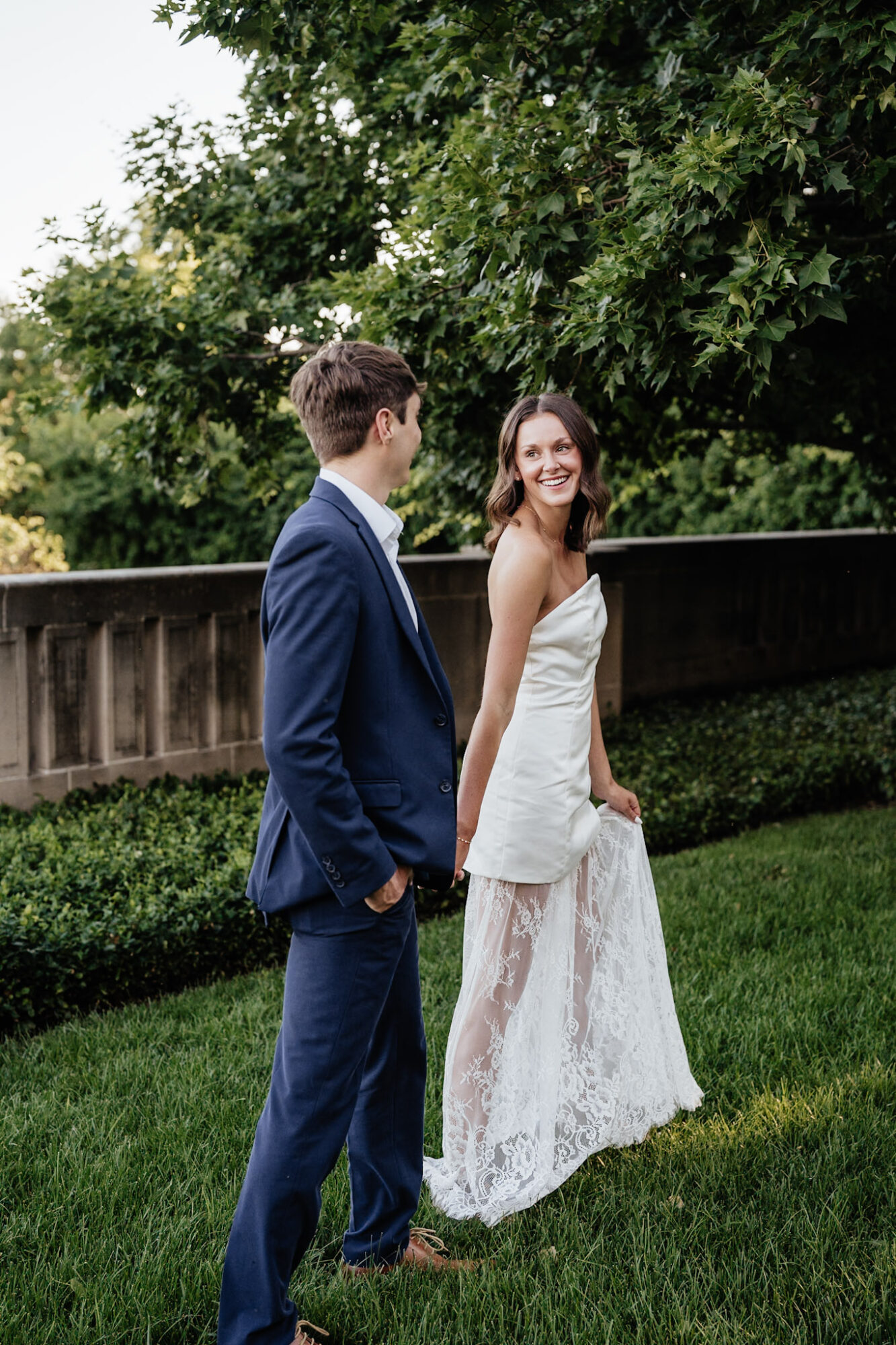 A smiling woman in a white dress and a man in a blue suit stand outdoors on grass with trees and a wooden fence behind them.