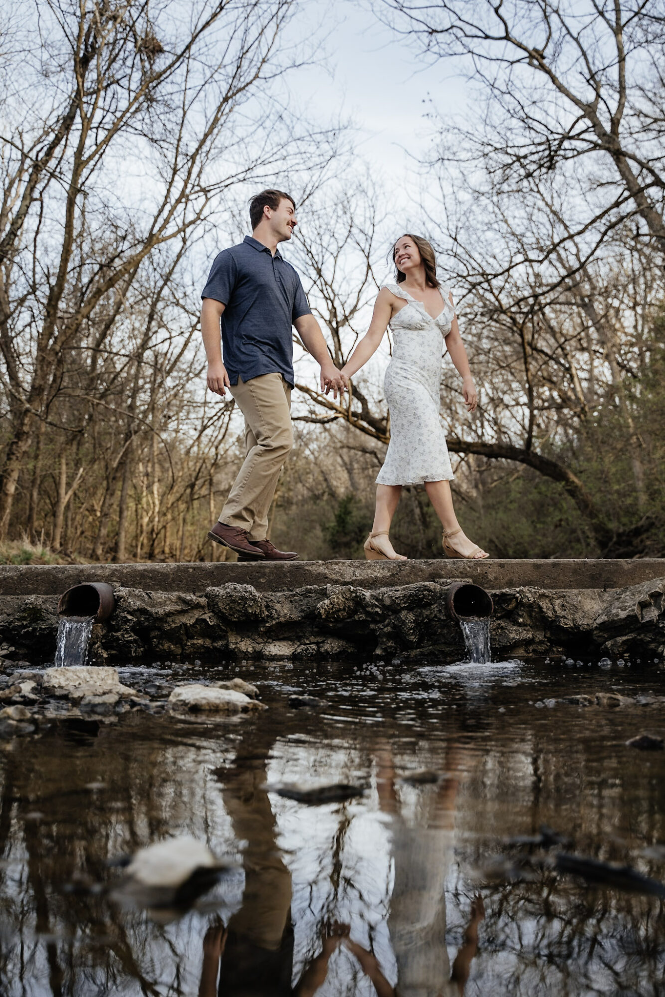 A couple holding hands walking on a log over a creek in a wooded area, reflected in the water below.