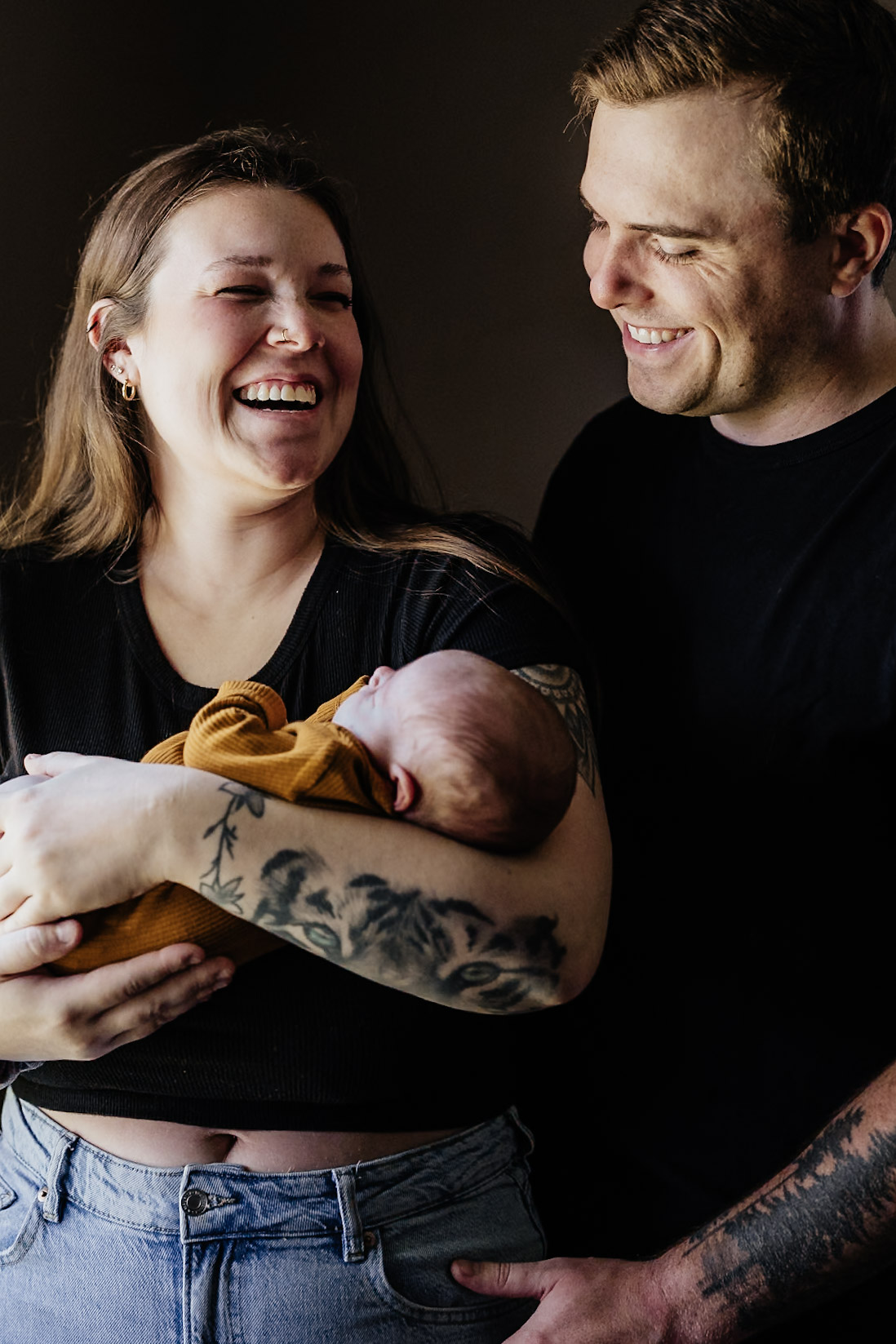 A woman holding a newborn baby, smiling, with a man standing beside her, smiling, in a dark background.