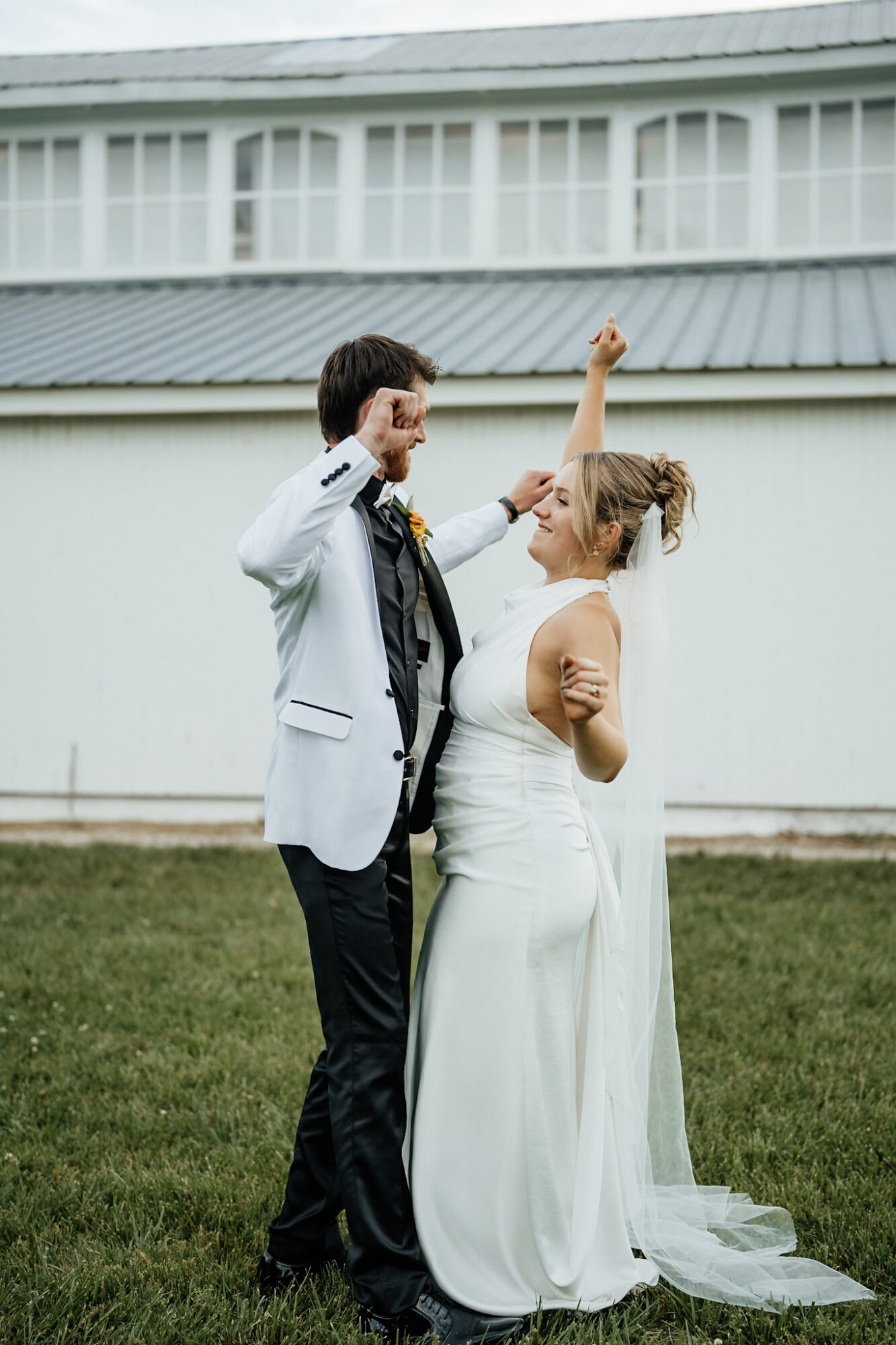A couple in wedding attire dancing outdoors on grass, with a building in the background.