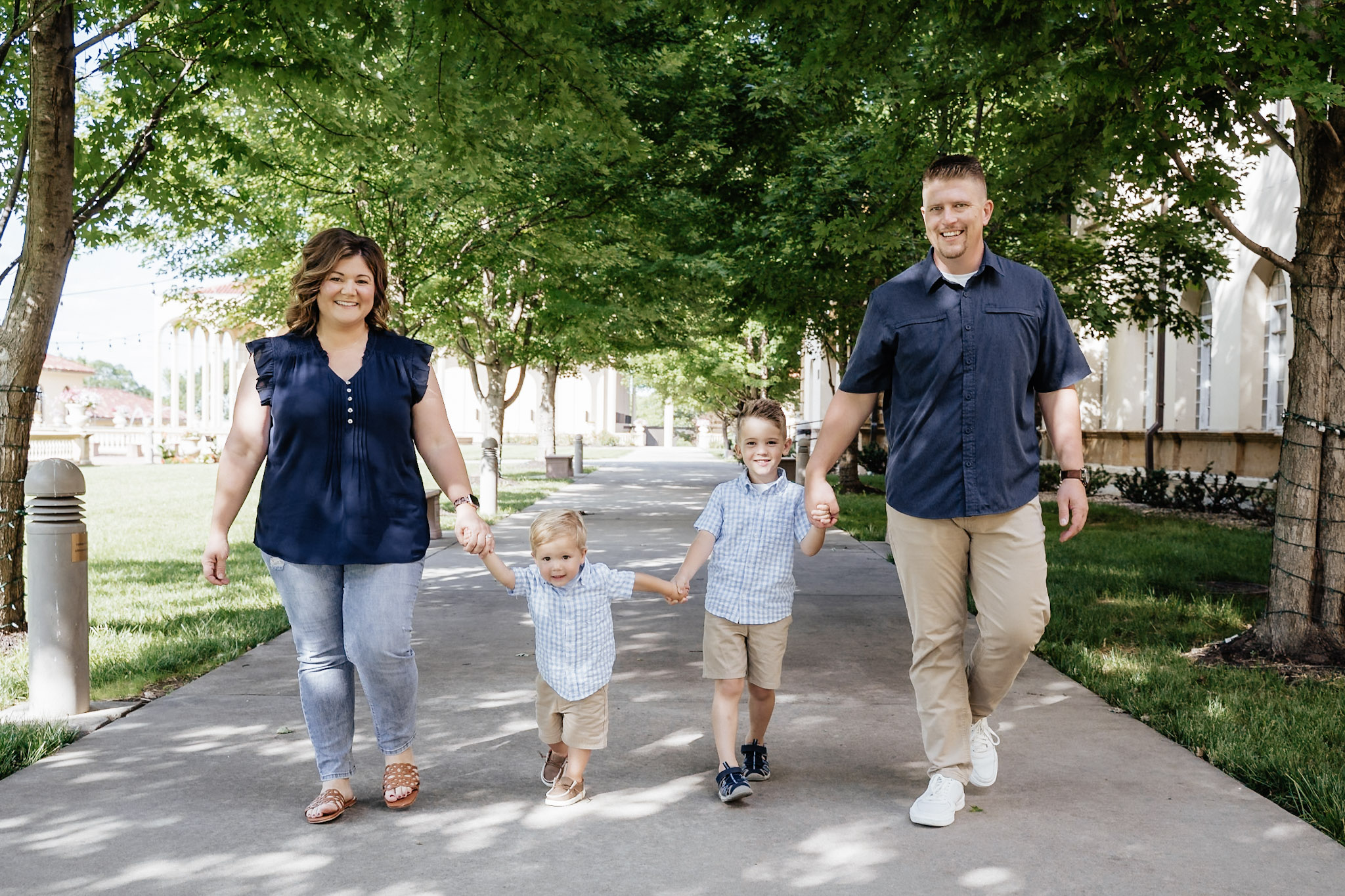 Family of four walking on a park path holding hands, trees and buildings in background, sunny day.