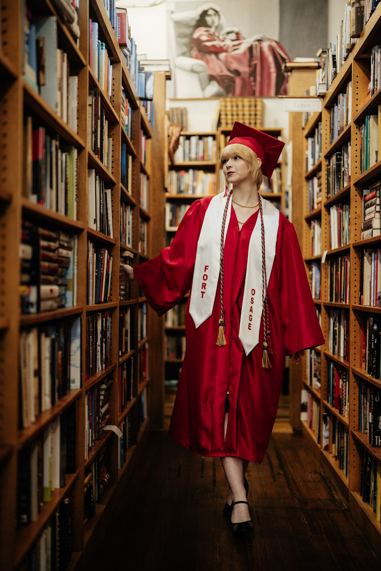 Woman in graduation gown and cap walking between bookshelves in a library, with a painting of a woman reading in the background.