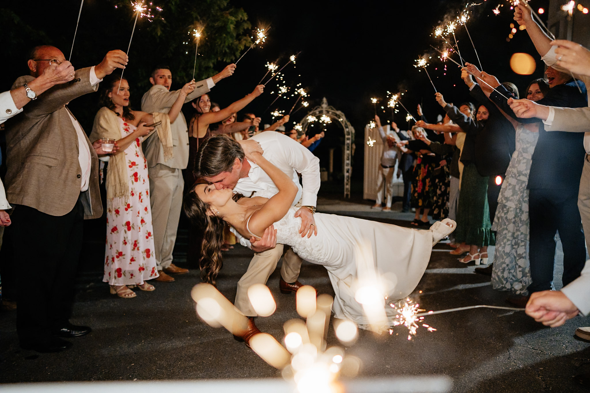 People celebrating at night, holding sparklers, with a woman in a white dress dancing in the center, candles on the ground, festive atmosphere.