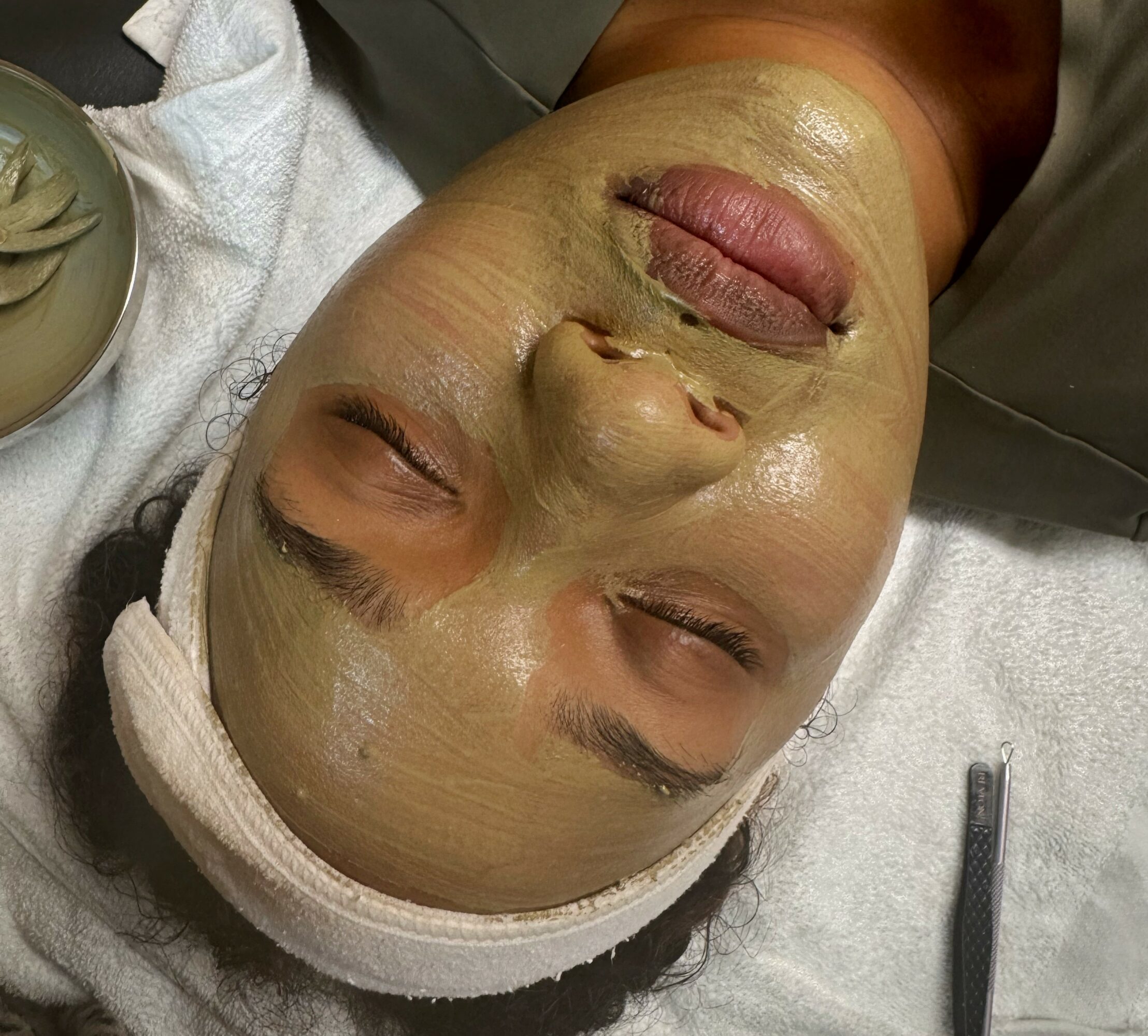 Person with a facial mask lying down with eyes closed, head wrapped in a towel, and a spa setting.