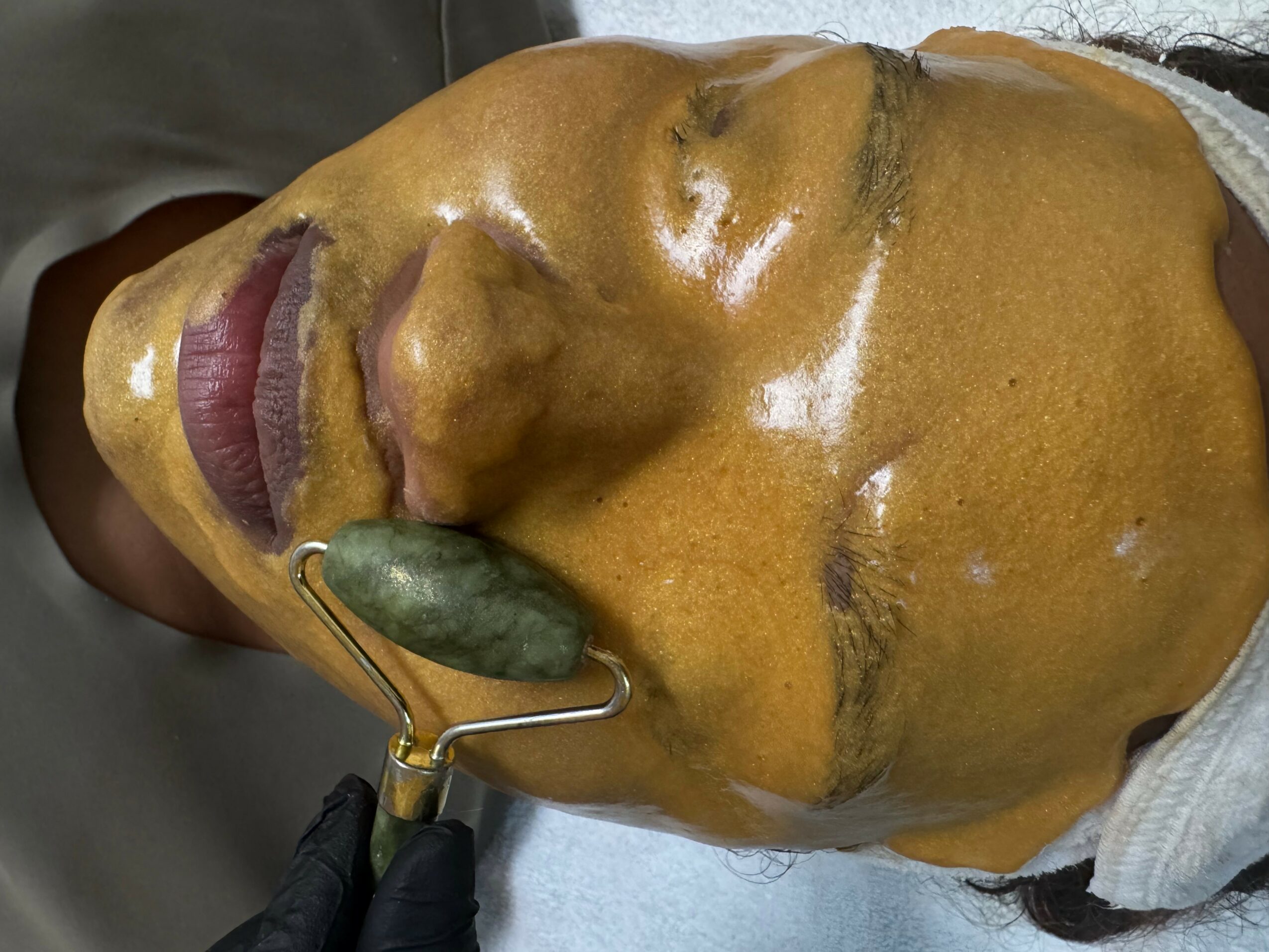 Person receiving facial treatment with a green jade roller, face covered in a yellow facial mask, lying down with eyes closed.