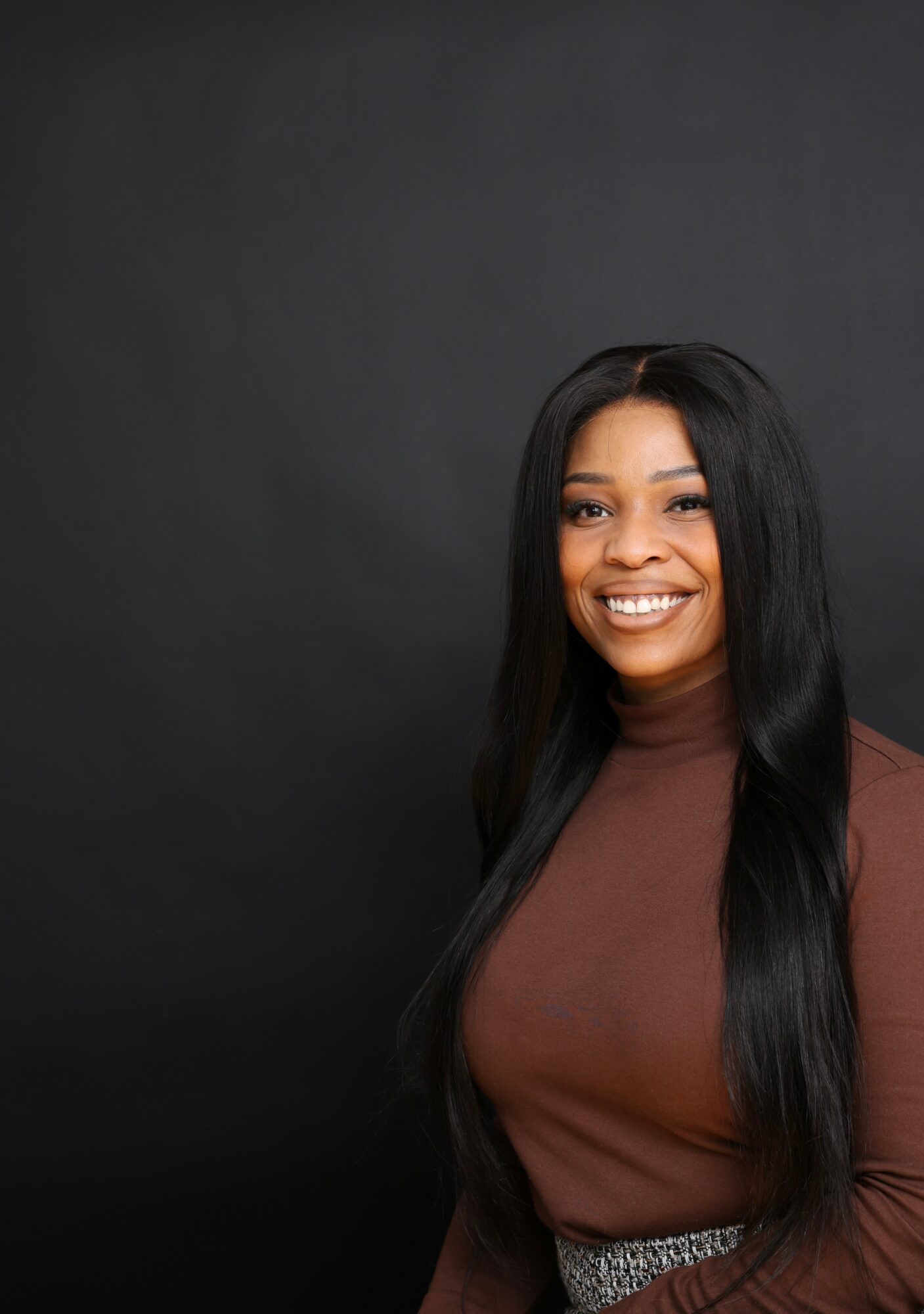 Smiling woman with long black hair wearing a brown top against a dark background.