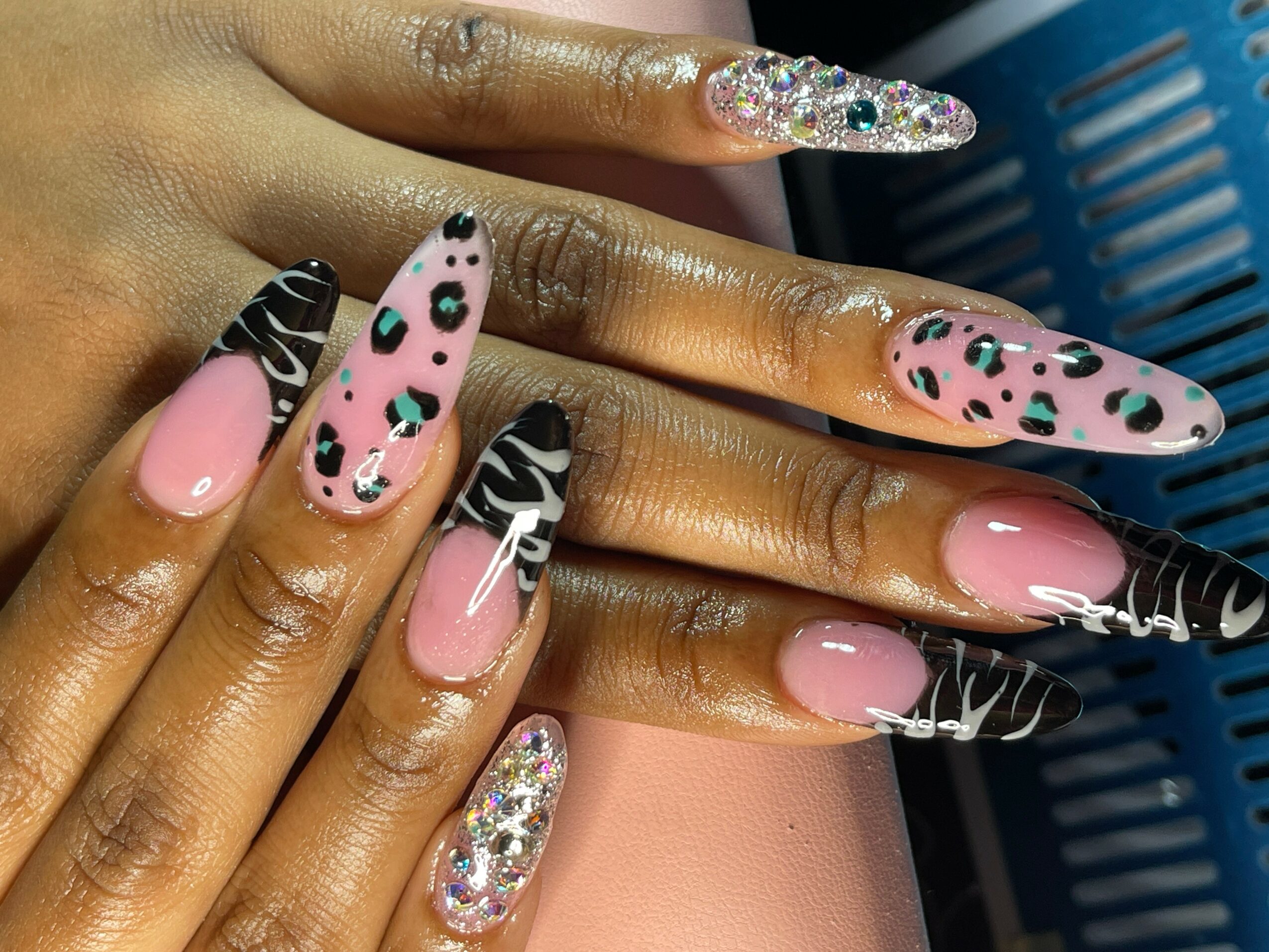 Close-up of hands with decorated nails, featuring pink, black, white, and glitter designs, against a blue background.