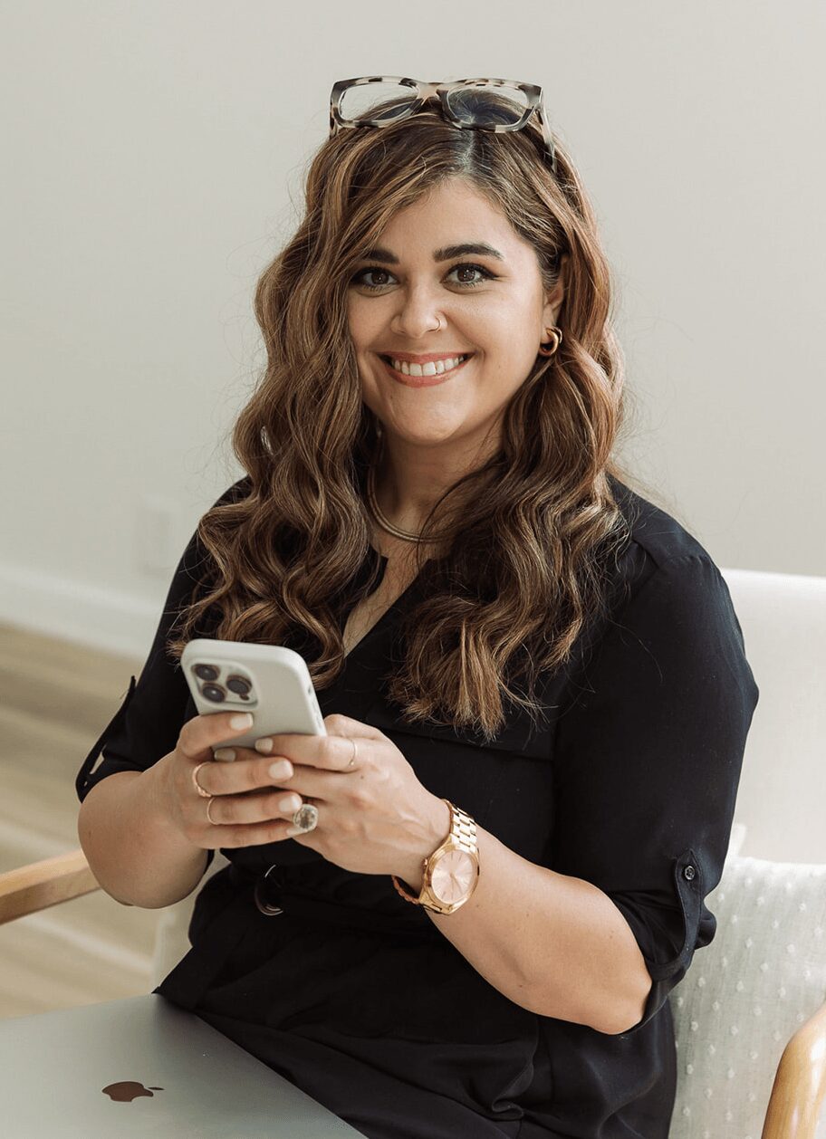 Young woman with wavy hair, glasses on head, smiling, holding a smartphone, wearing a black top, sitting indoors.