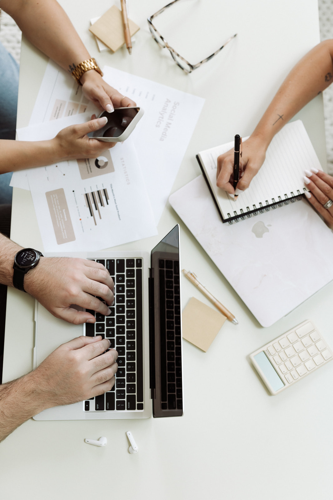 People working at a desk with a laptop, smartphone, notebook, and documents, with hands visible. Word count: 20.