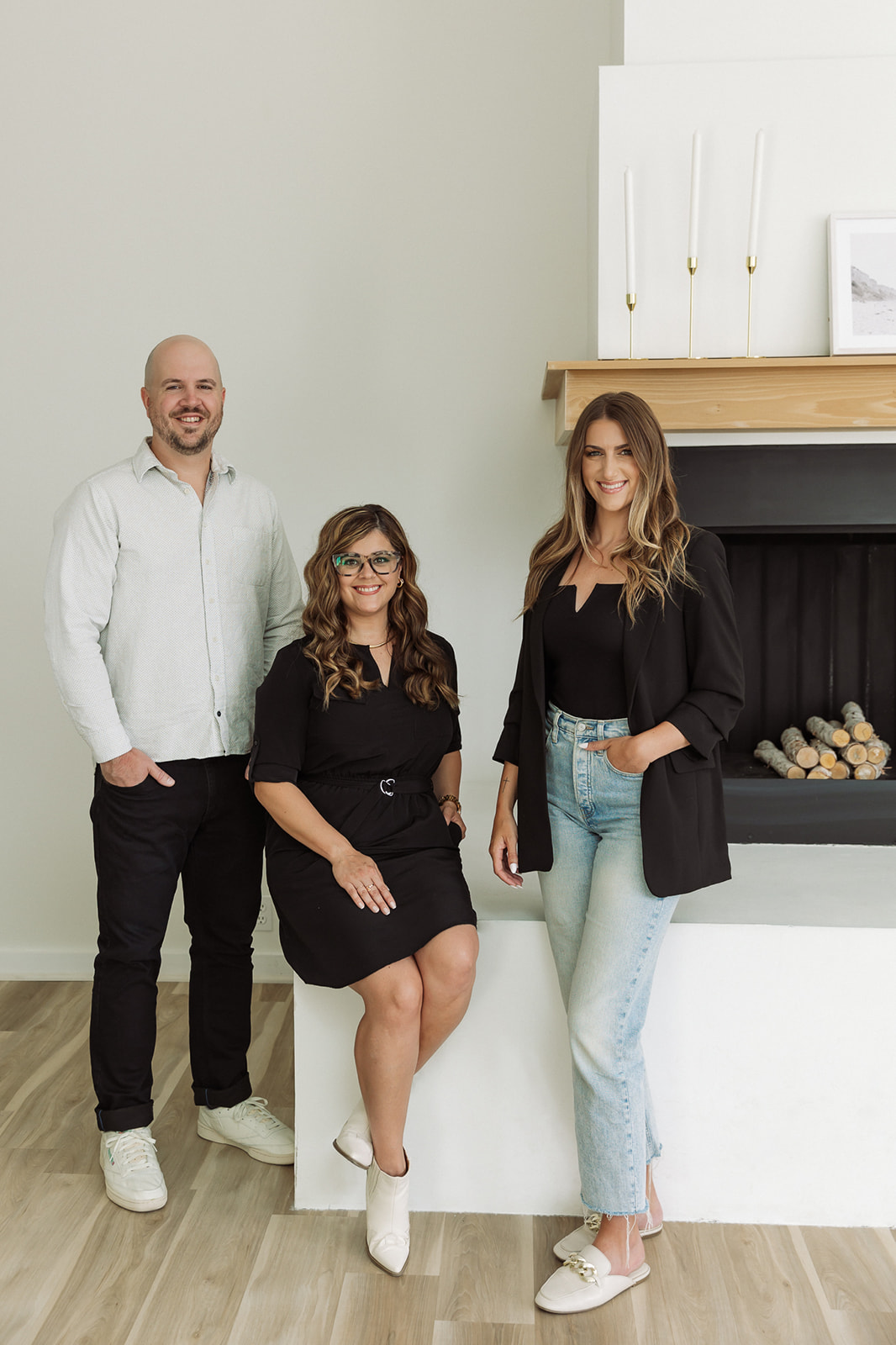 Three people, two women and one man, standing and sitting in a modern room with a fireplace and white cabinets, smiling.
