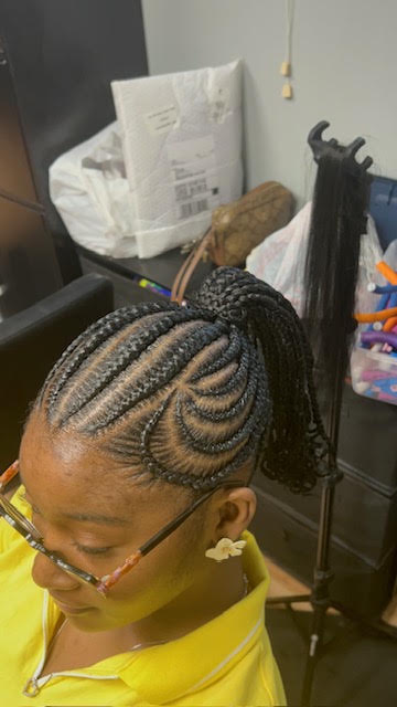 Person with braided hairstyle sitting in a room with various items in the background.