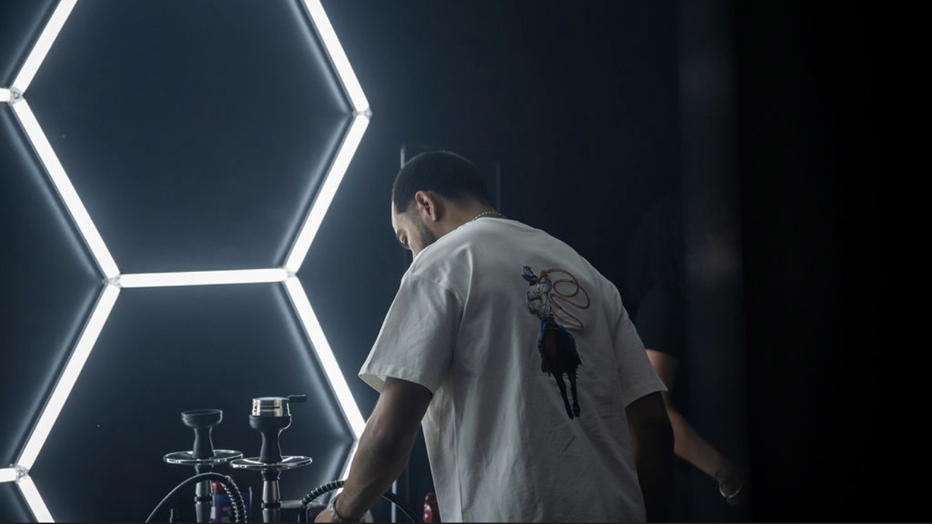 Person with short hair wearing a white shirt with a black deer design, standing near hexagonal light panels, facing away.