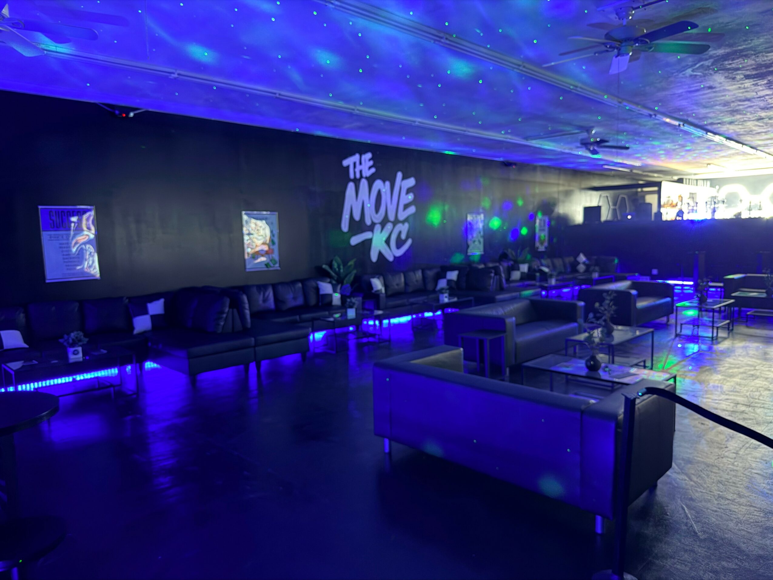 Dark indoor space with illuminated seating, a wall with screens, and a projection reading 'The Move KC' with blue lighting.