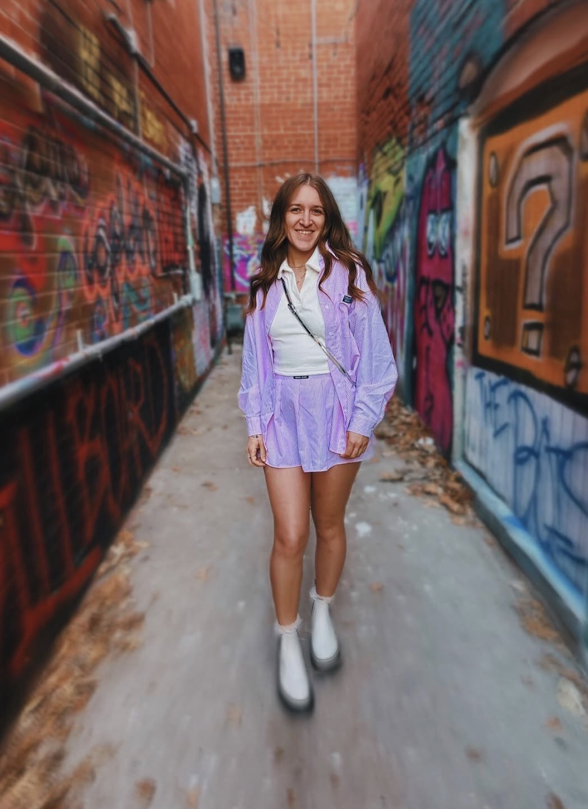Young woman walking in graffiti-covered alley, smiling, wearing purple jacket, shorts, and white boots.