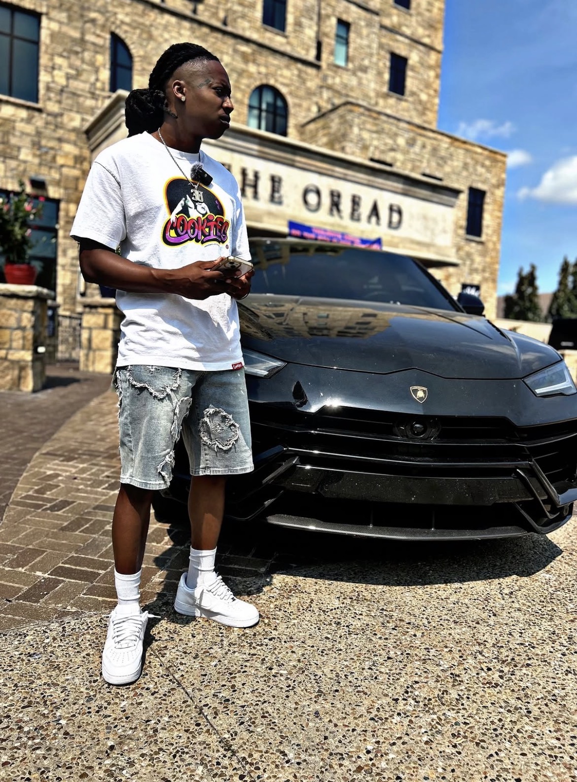 Young man with dreadlocks in a white t-shirt, denim shorts, and sneakers stands next to a black Lamborghini in front of a building with a sign.