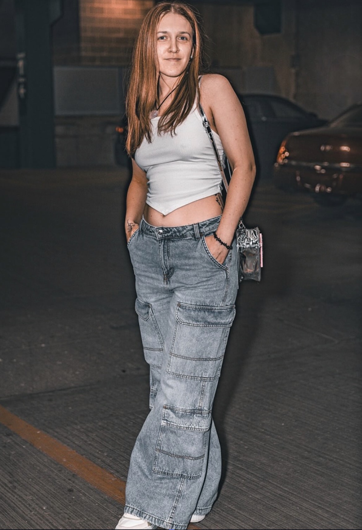 Young woman with long hair in a white crop top and baggy jeans standing outdoors at night, hands in pockets.