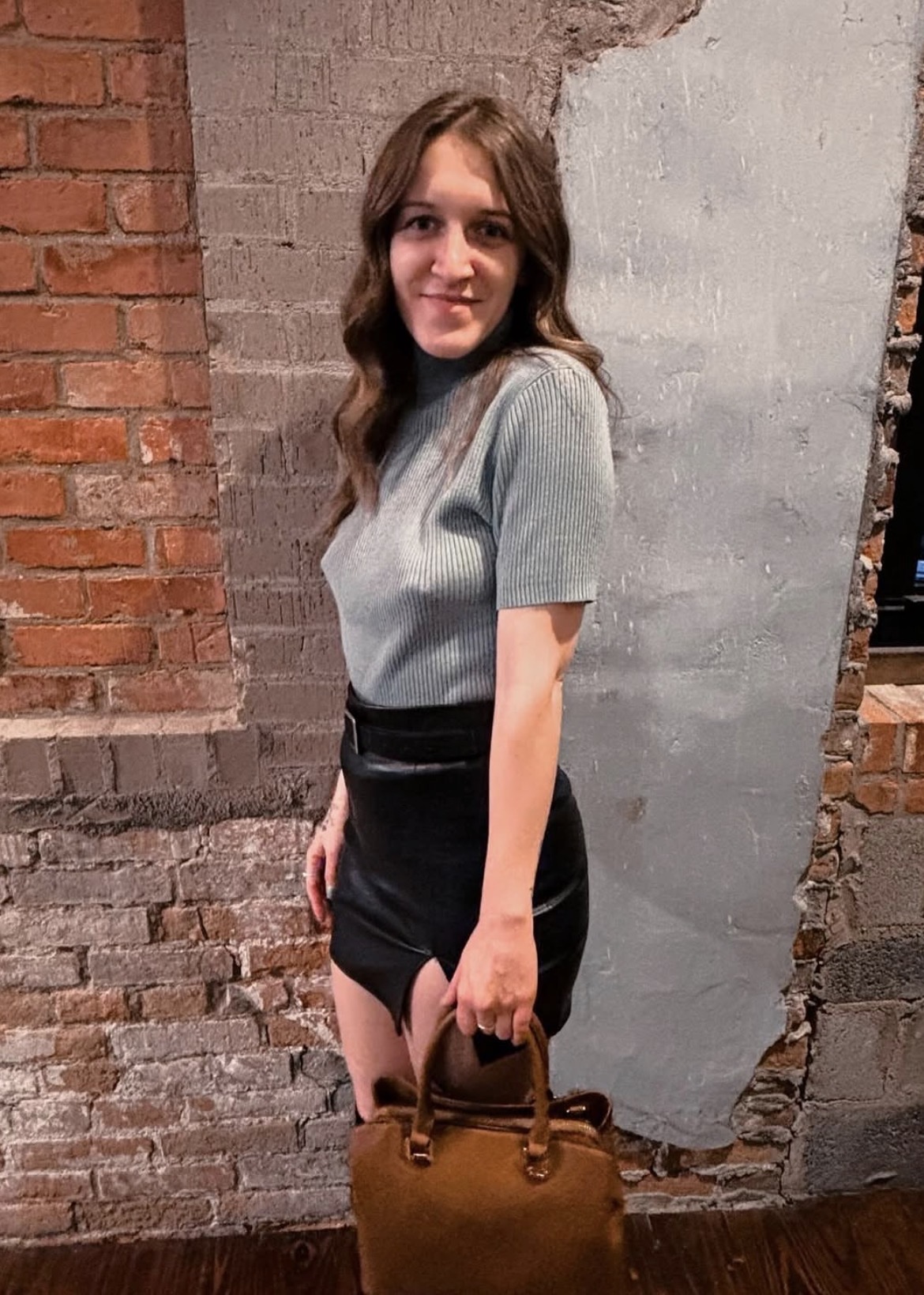 Young woman with wavy brown hair in a gray top and black skirt holding a brown handbag, standing against a brick wall.