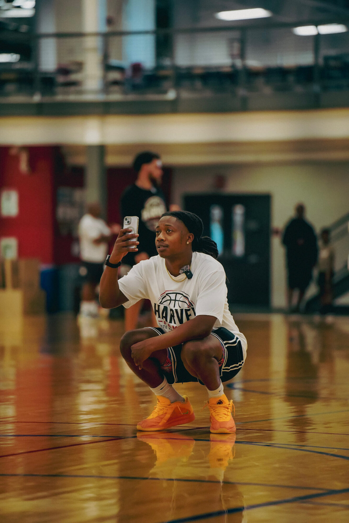 Young basketball player crouching on court, holding a ball, taking a selfie, with people and gym background.