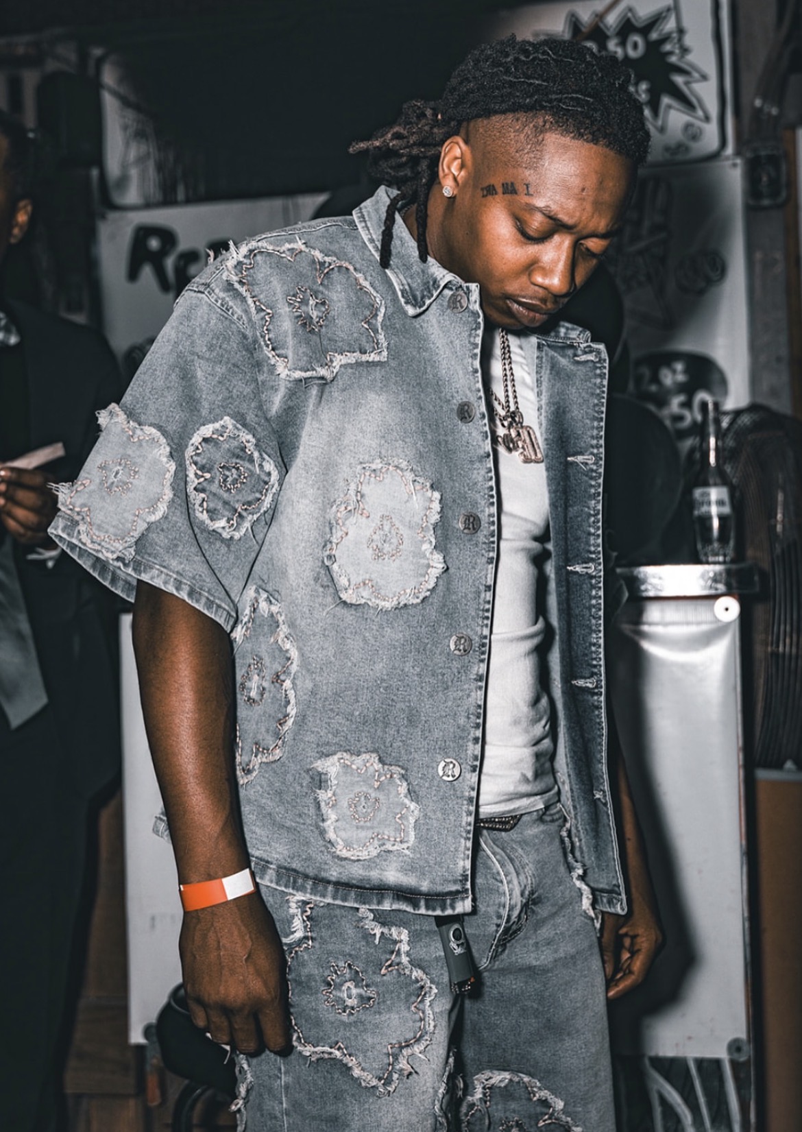 Man with dreadlocks wearing a distressed denim jacket and jeans, standing in a dimly lit indoor setting.